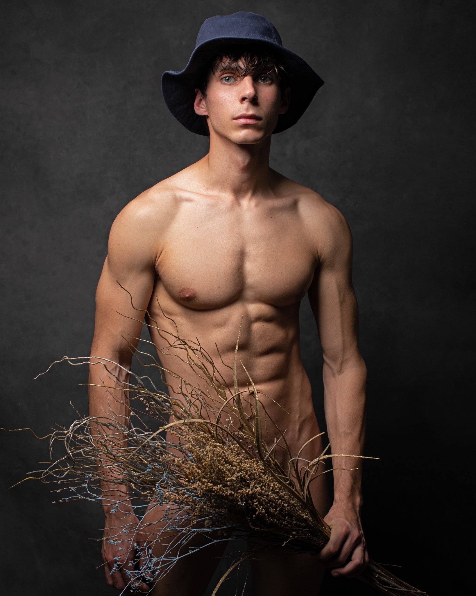 A shirtless young man with a lean, muscular physique wearing a dark wide-brimmed hat. He is holding a large bouquet of dried, brown, and curly stems in his right hand, standing against a plain dark background.