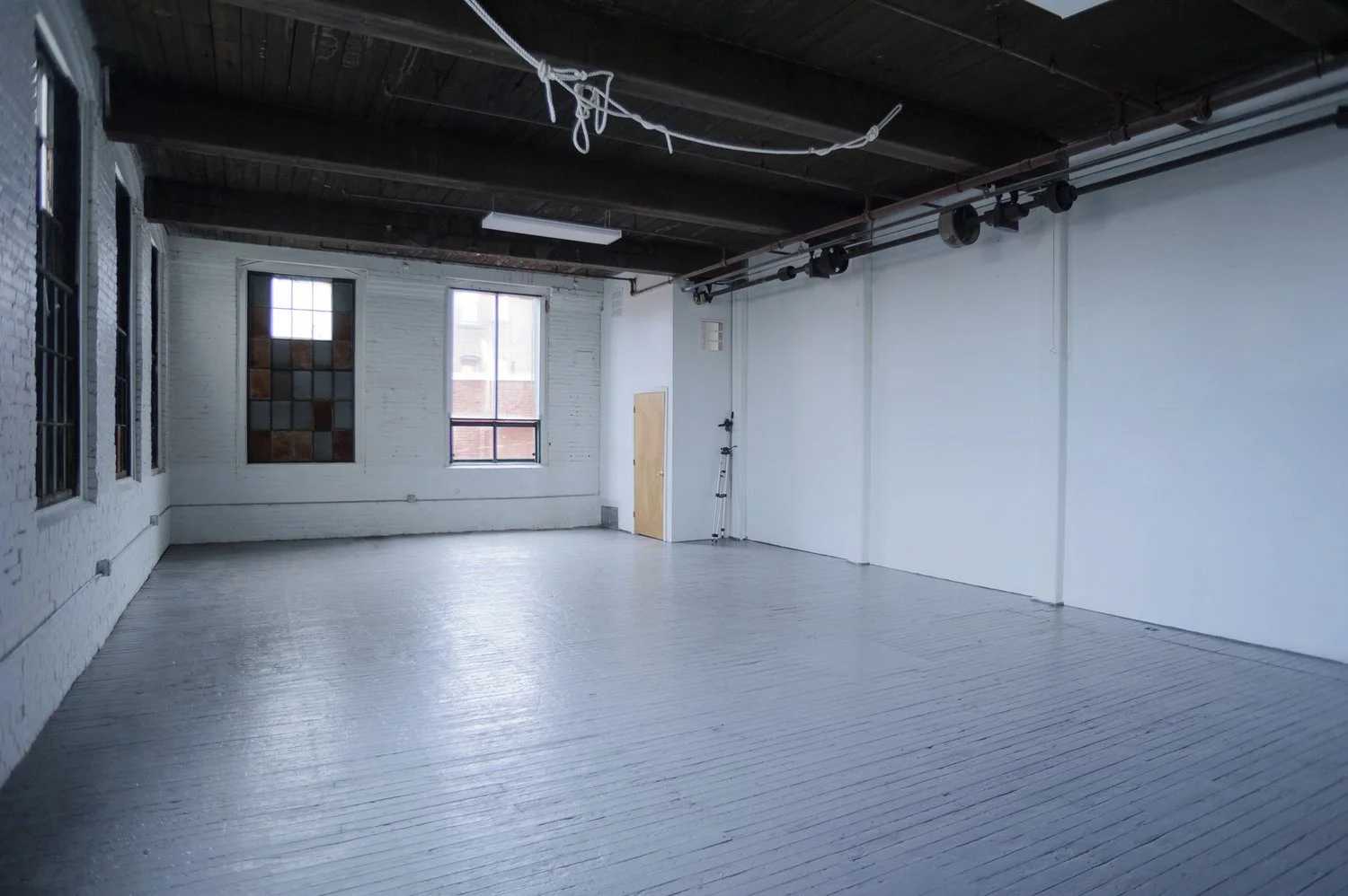 Empty industrial-style room with white wooden floors, white brick and painted walls, large windows, and a dark exposed ceiling with some pipe and wiring