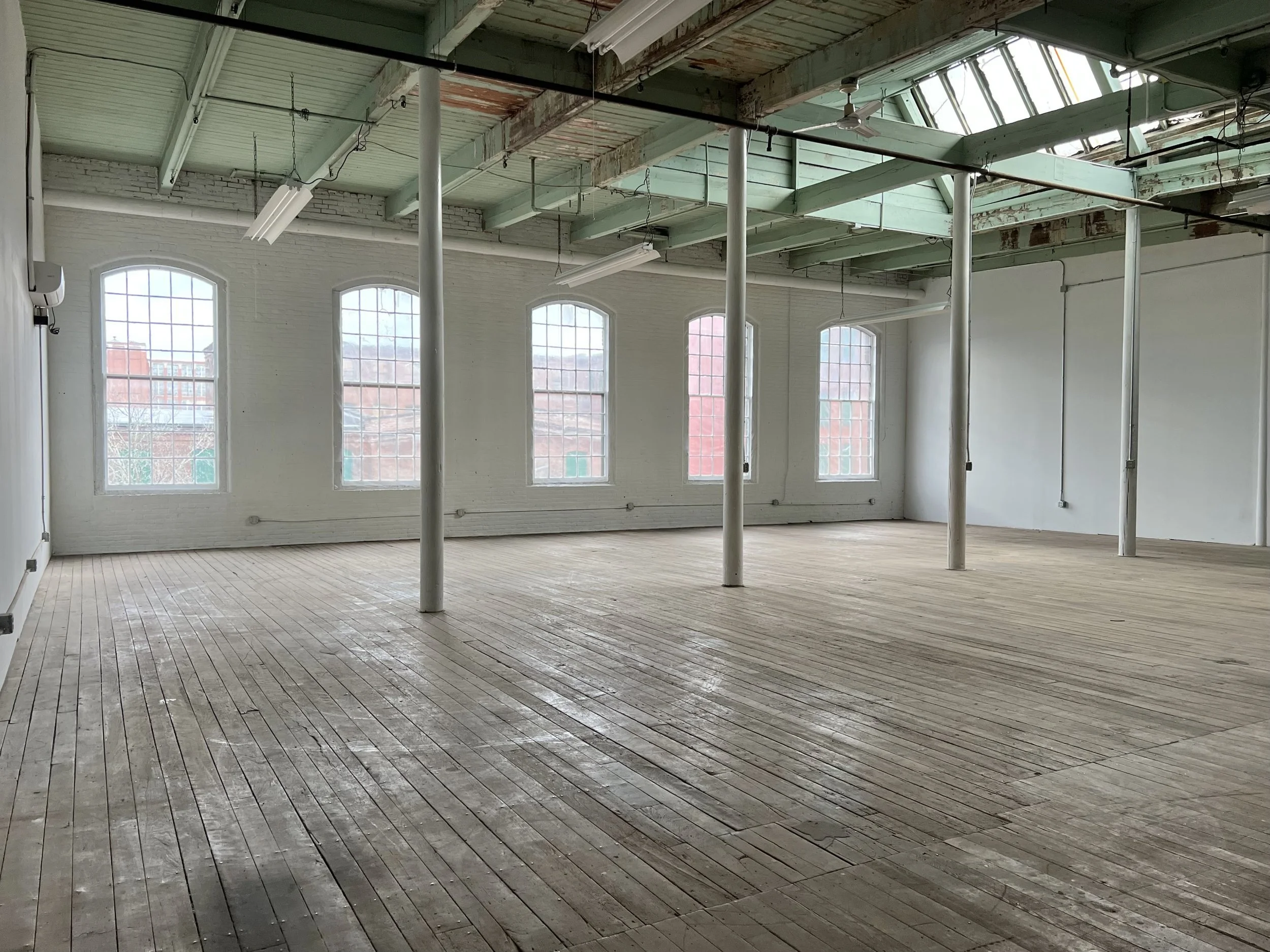 Empty industrial-style room with large windows, wooden floor, and exposed ceiling beams and pipes.