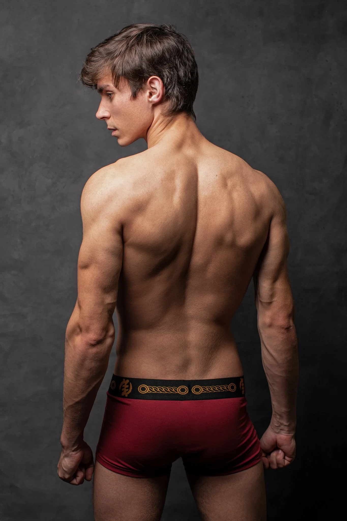 A shirtless young man with short brown hair, facing away from the camera, wearing red and black underwear, standing against a dark textured background.