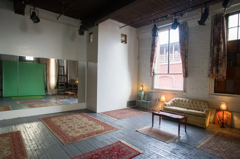 A spacious studio with wooden floors, large window with floral curtains, a beige tufted sofa, two small lamps on side tables, a green vintage chair, and multiple Persian rugs. There is a wall with a mirror and a green screen or backdrop, and a ceiling with track lighting.