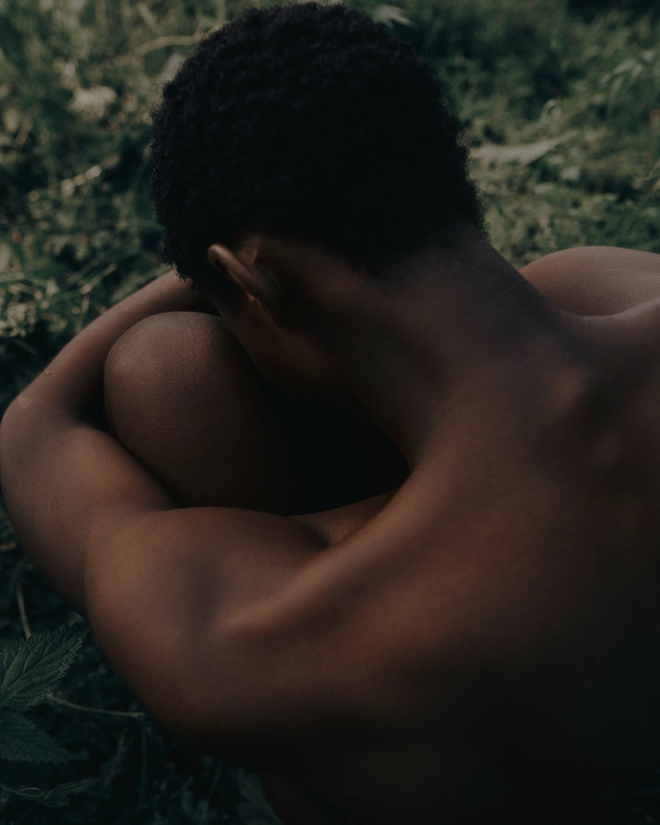 A person with dark skin and short curly hair sitting outdoors, hugging their knees with their head resting on their arms, surrounded by green foliage.