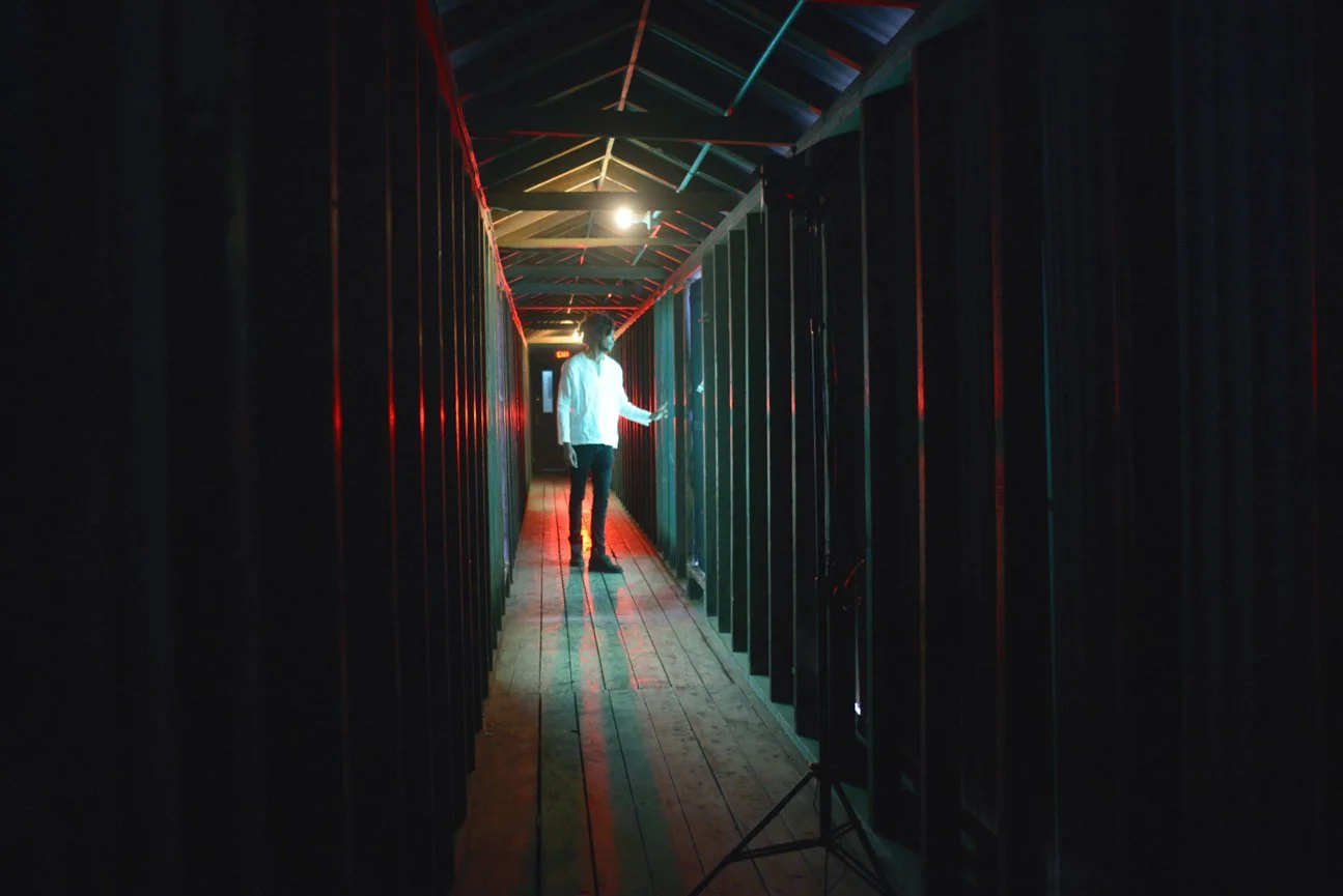 A person standing in a dark, narrow hallway illuminated by colorful lights, with a wooden floor and a peaked ceiling.