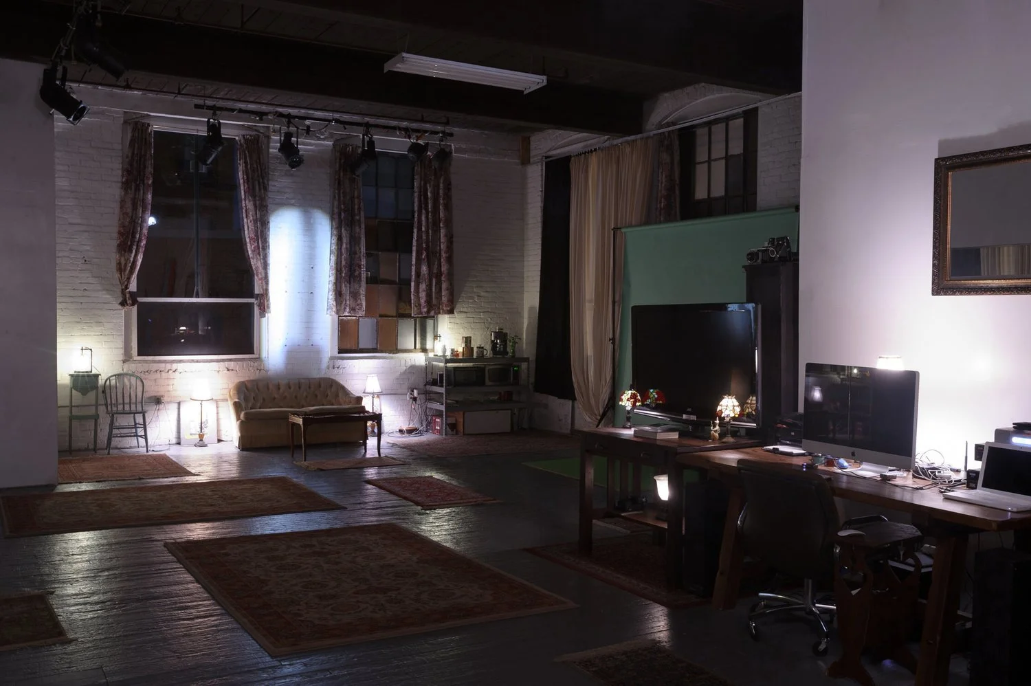 A dimly lit living room with a sofa, chairs, rugs, and a workspace with two computer monitors, a window with curtains, and a large TV on a stand.