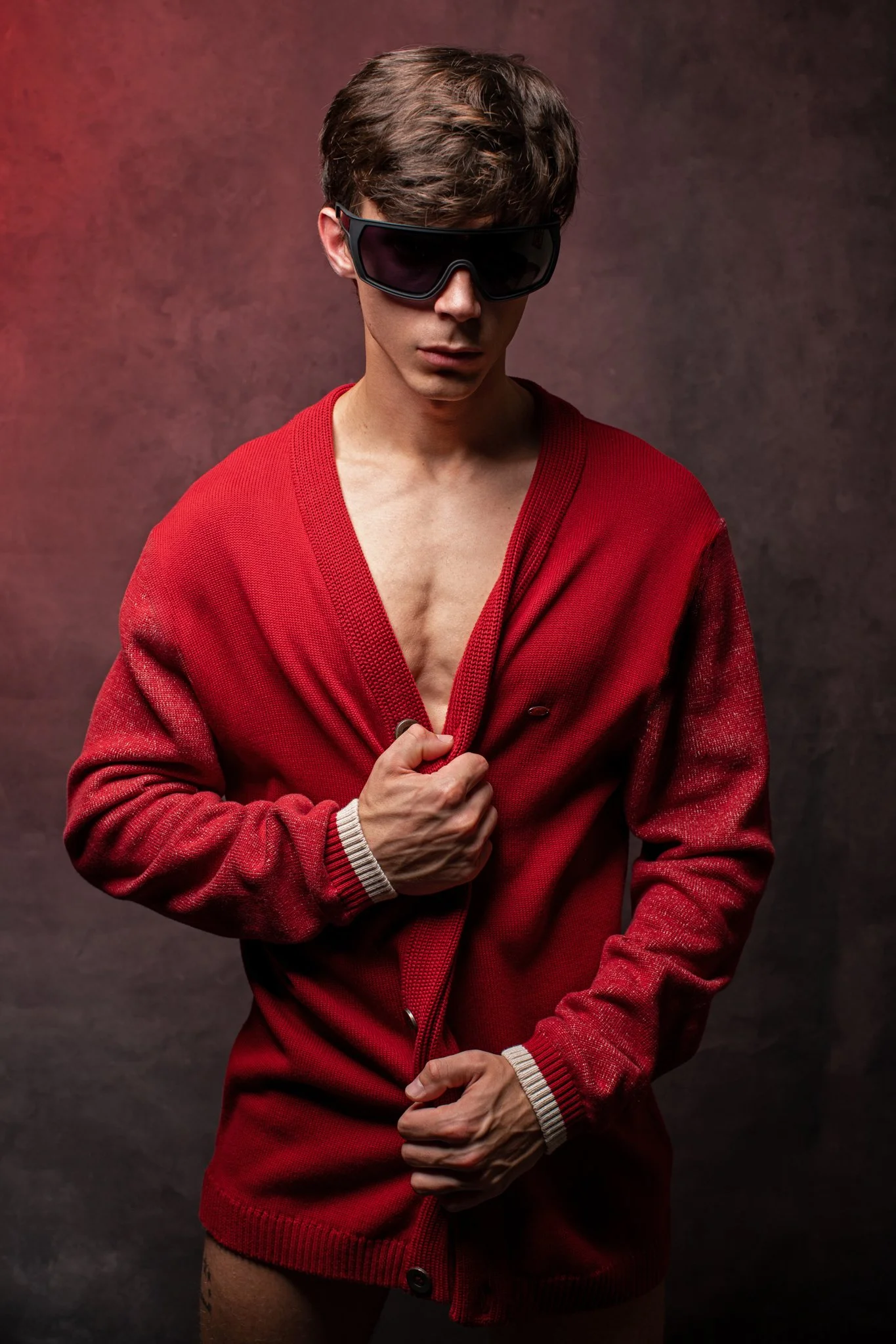 A young man wearing large black sunglasses and a red cardigan, partially unbuttoned, revealing his chest, poses against a dark background.