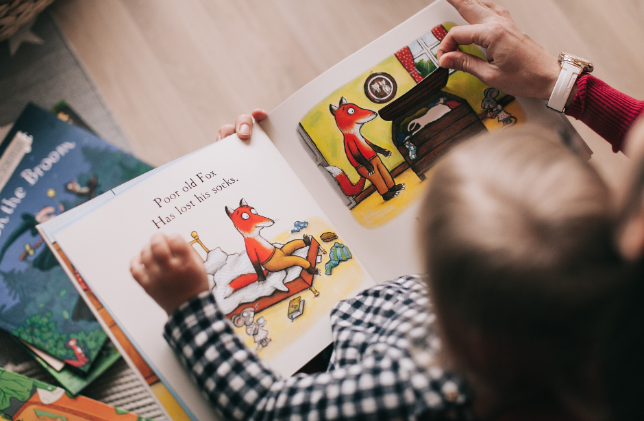 A child reading a colorful children's book about a fox. The book shows a fox sitting on a bed with the text 'Poor old Fox Has lost his socks.' The child is turning the page to a scene with the fox in a different room, sitting on a chair. There are other books around, and the child has a watch on their wrist.