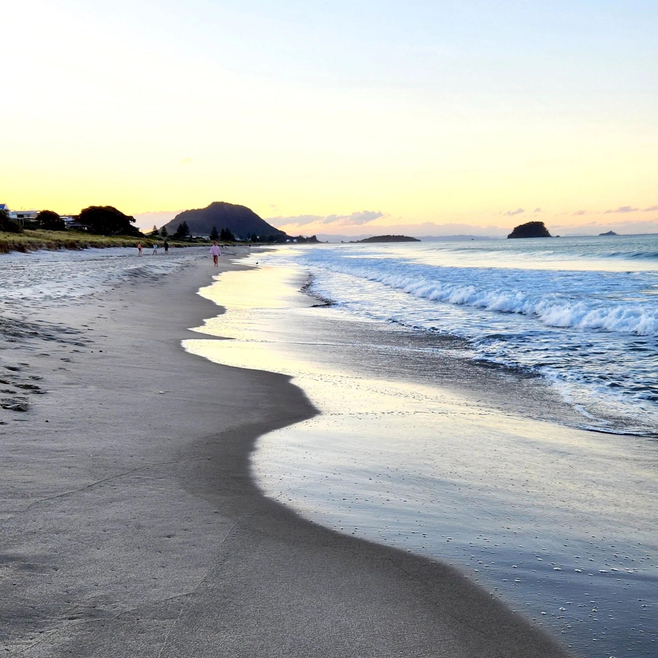 A sandy beach during sunset with gentle waves washing ashore and a few people walking along the beach, with hills and small islands in the distance.