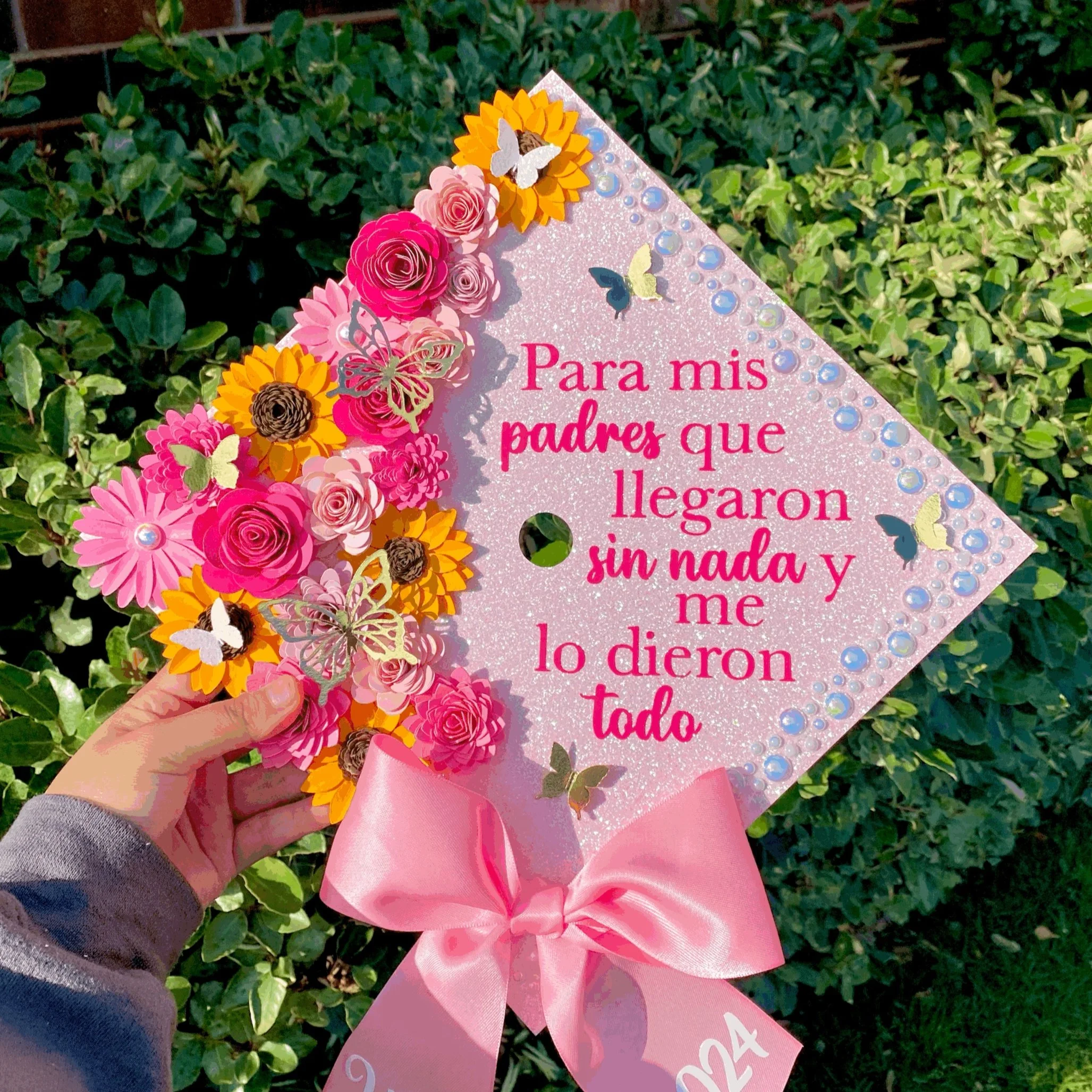 Decorative graduation cap decorated with pink, yellow, and brown flowers and butterflies, with a pink ribbon bow, featuring a Spanish message for parents indicating achievement without material wealth.
