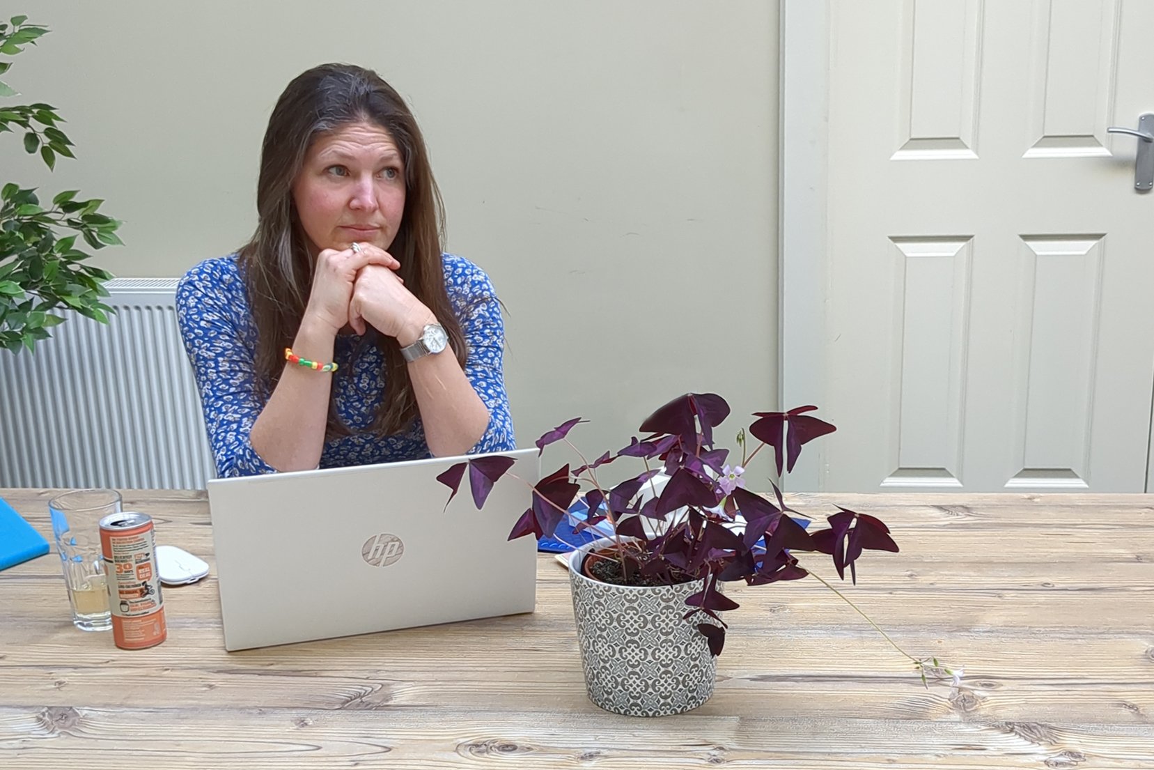 A woman sitting at a wooden table with a laptop, a glass of waterand a blue notebook. There is a potted purple plant in front of her and a large green plant to her left. She has long brown hair and is wearing a blue patterned bracelet. She is resting her chin on her folded hands and looking to her left.