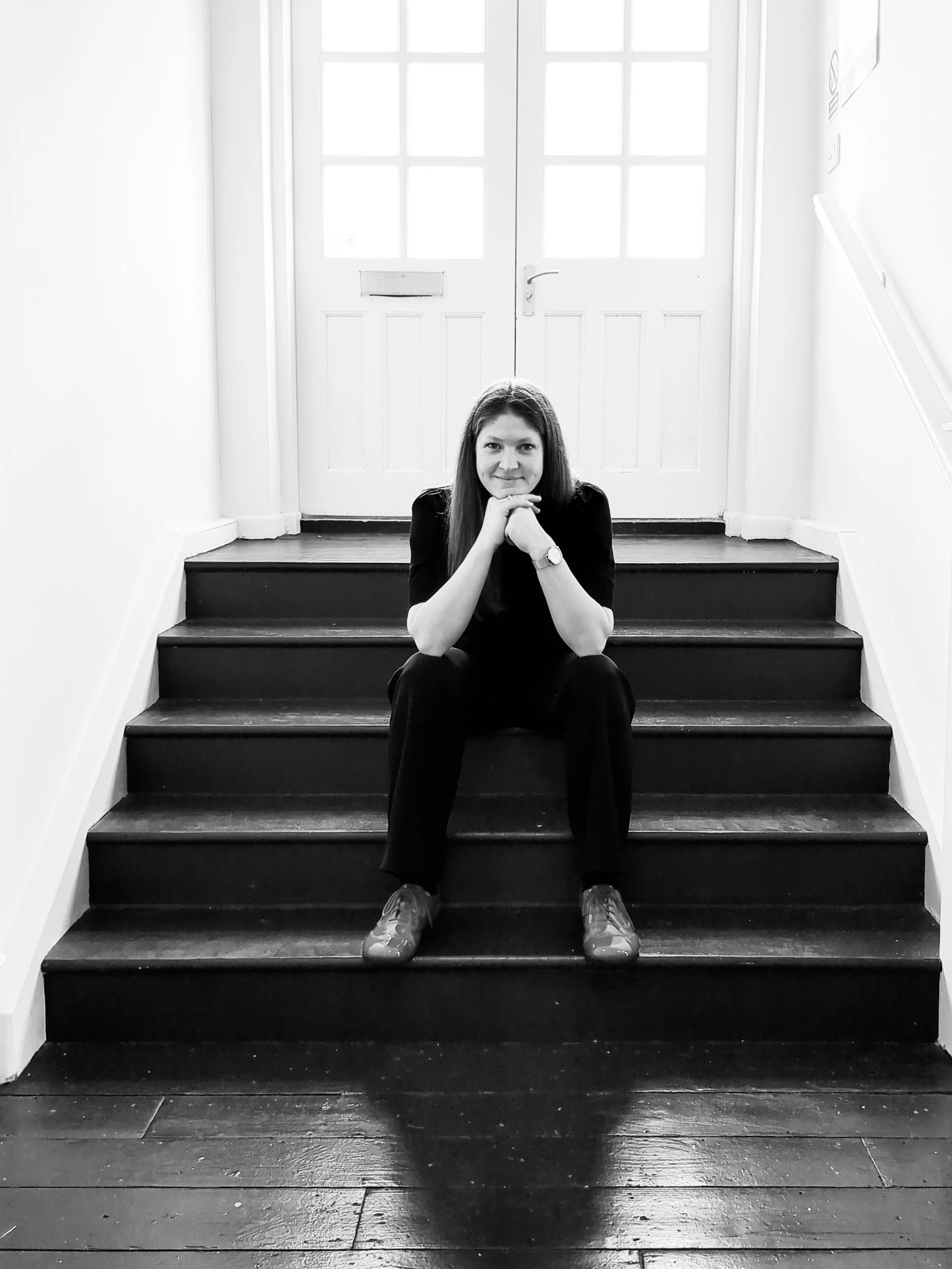 A woman, Yvi (Yvonne) Guerineau, sitting on wooden stairs in front of a set of white double doors with glass panes, smiling and resting her chin on her hands.