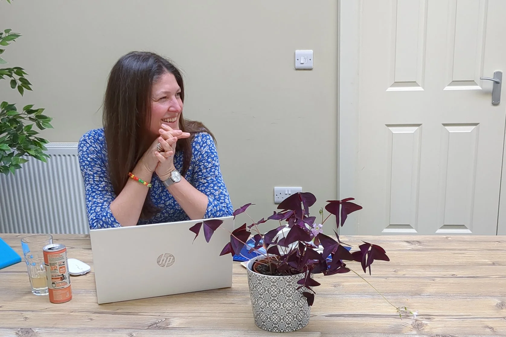 A woman sitting at a wooden table in an office, smiling and looking to her left, with a laptop, a glass, and a small can on the table, and plants in the foreground.