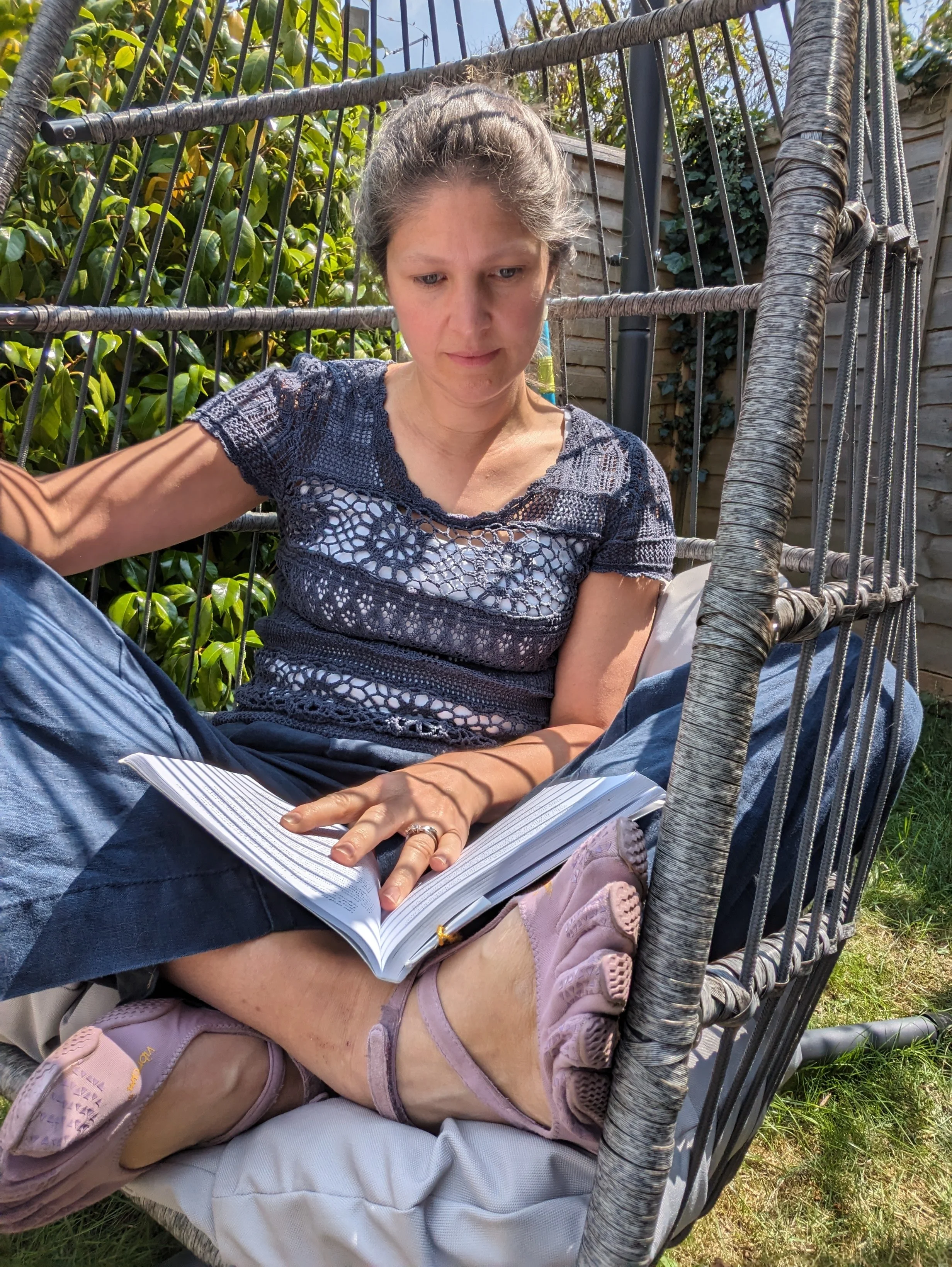 A woman , Yvi (Yvonne) Guerineau, sitting in a hanging garden chair outdoors during the day, engrossed in a book. She is wearing a navy blue lace top, jeans, and pink barefoot shoes. There is greenery and a wooden fence in the background.