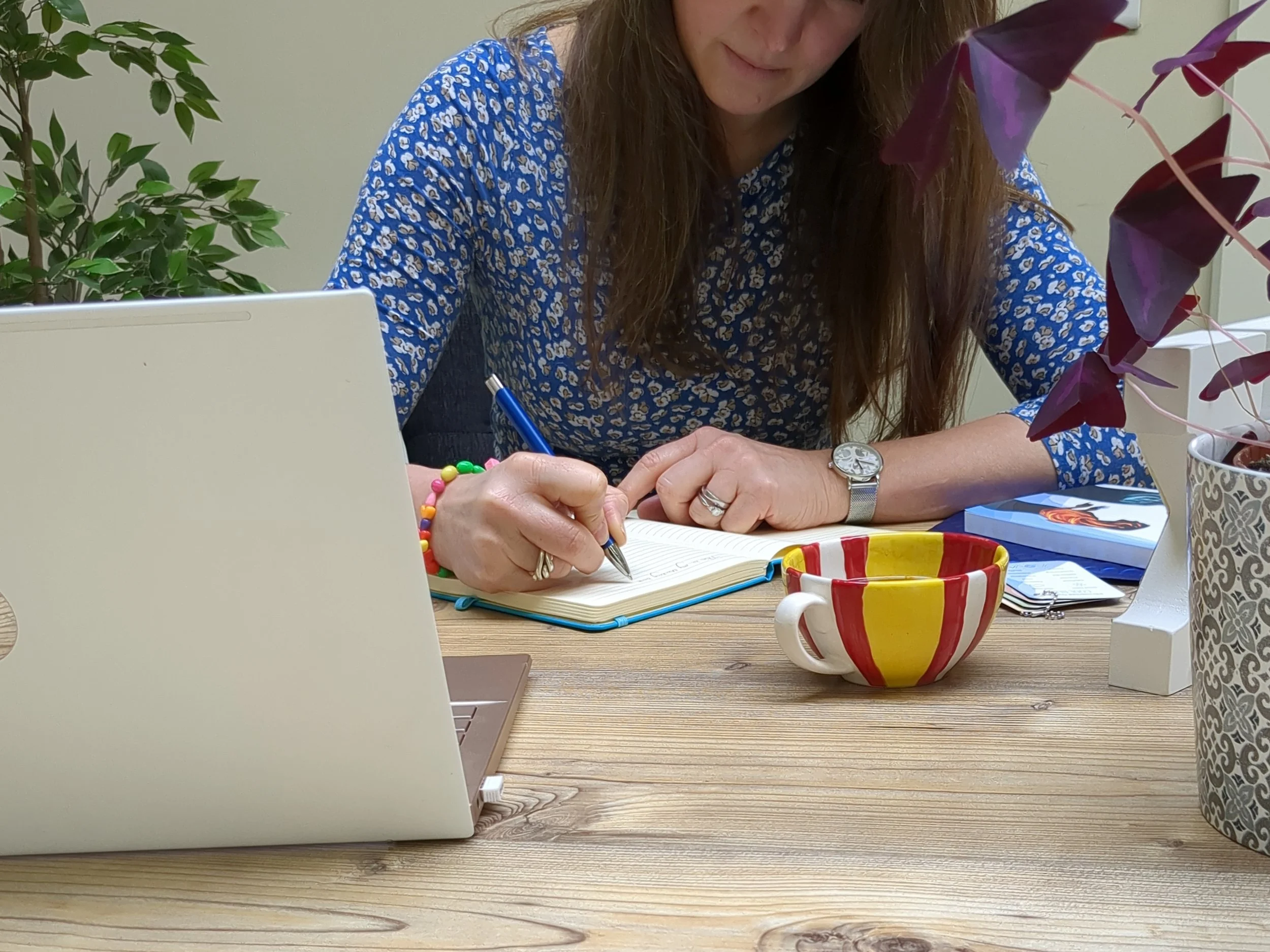 A woman, Yvi (Yvonne) Guerineau, with long brown hair, wearing a blue patterned shirt, sitting at a desk with various items, writing in a notebook. There are plants and a colorful mug on the desk.