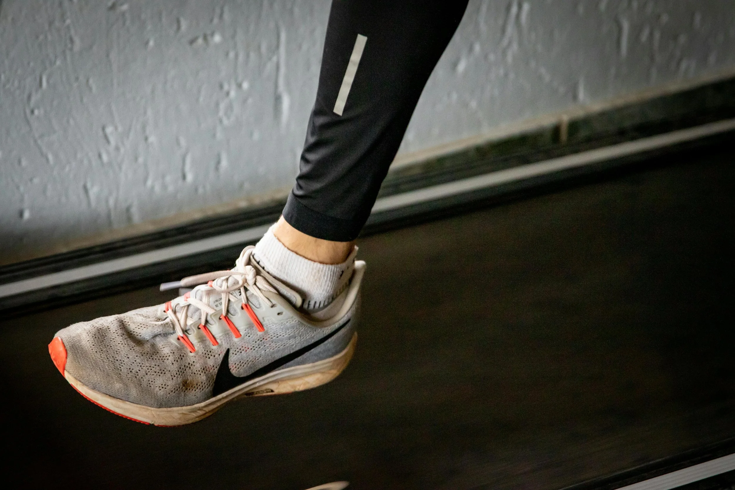Close-up of a person running on a treadmill, wearing a gray Nike running shoe, black athletic leggings, and a white ankle sock.