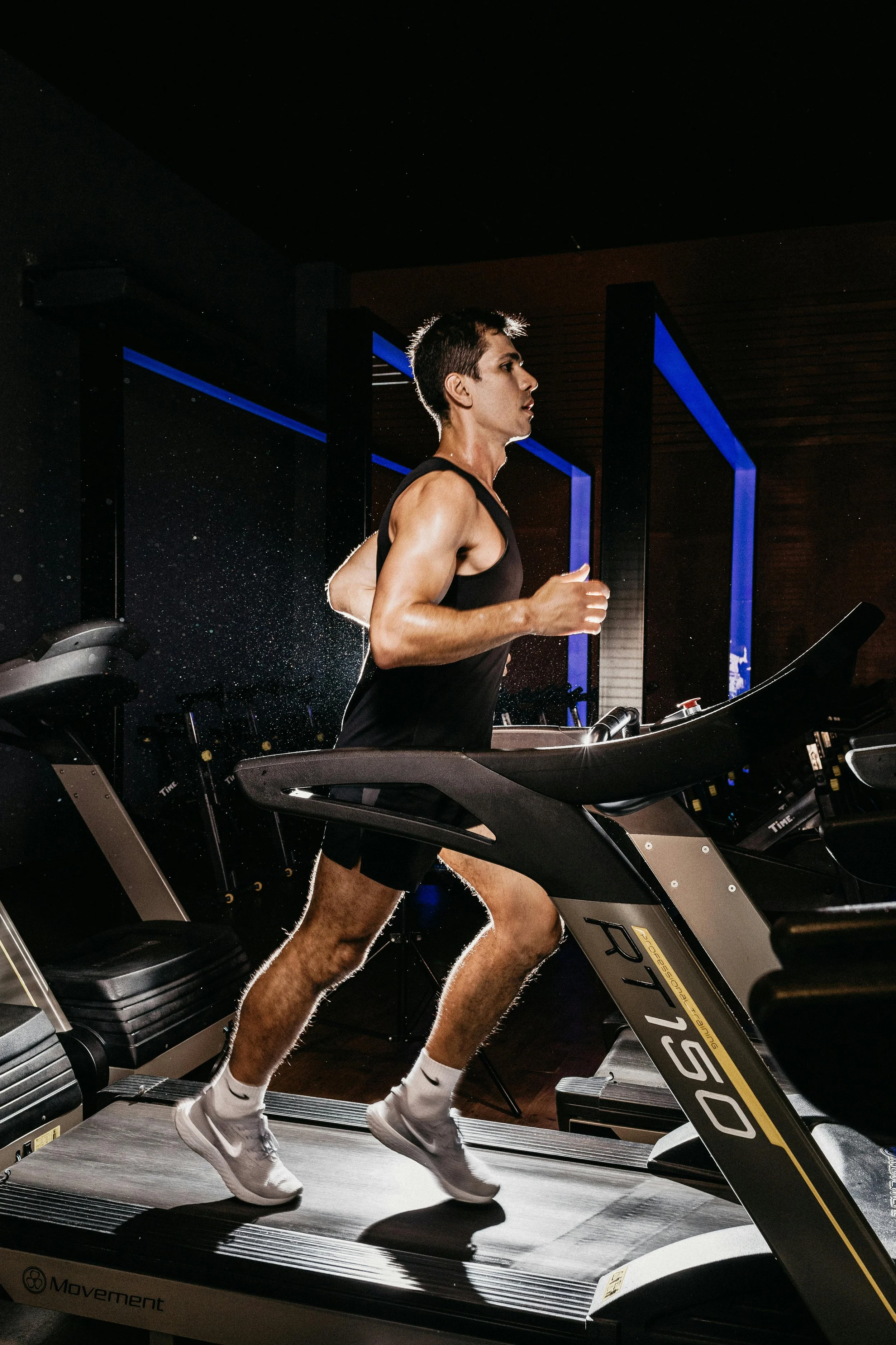 A man running on a treadmill in a dark gym with blue accent lighting.