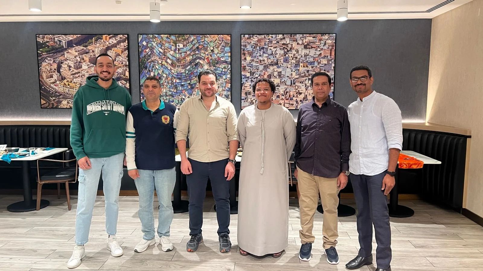 Seven men standing side by side in a restaurant or cafe with art photographs on the wall behind them.