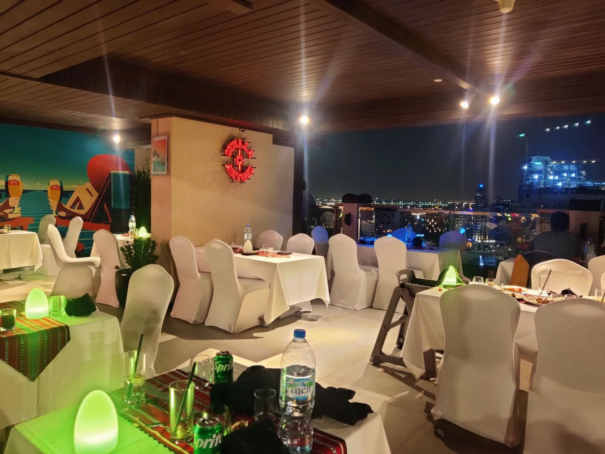 Indoor rooftop restaurant at night with white covered chairs and tables set with glasses, bottles, and tableware. City skyline with lights visible through large windows in the background. A Stella Artois neon sign on the wall, colorful wall mural, and ambient lighting.