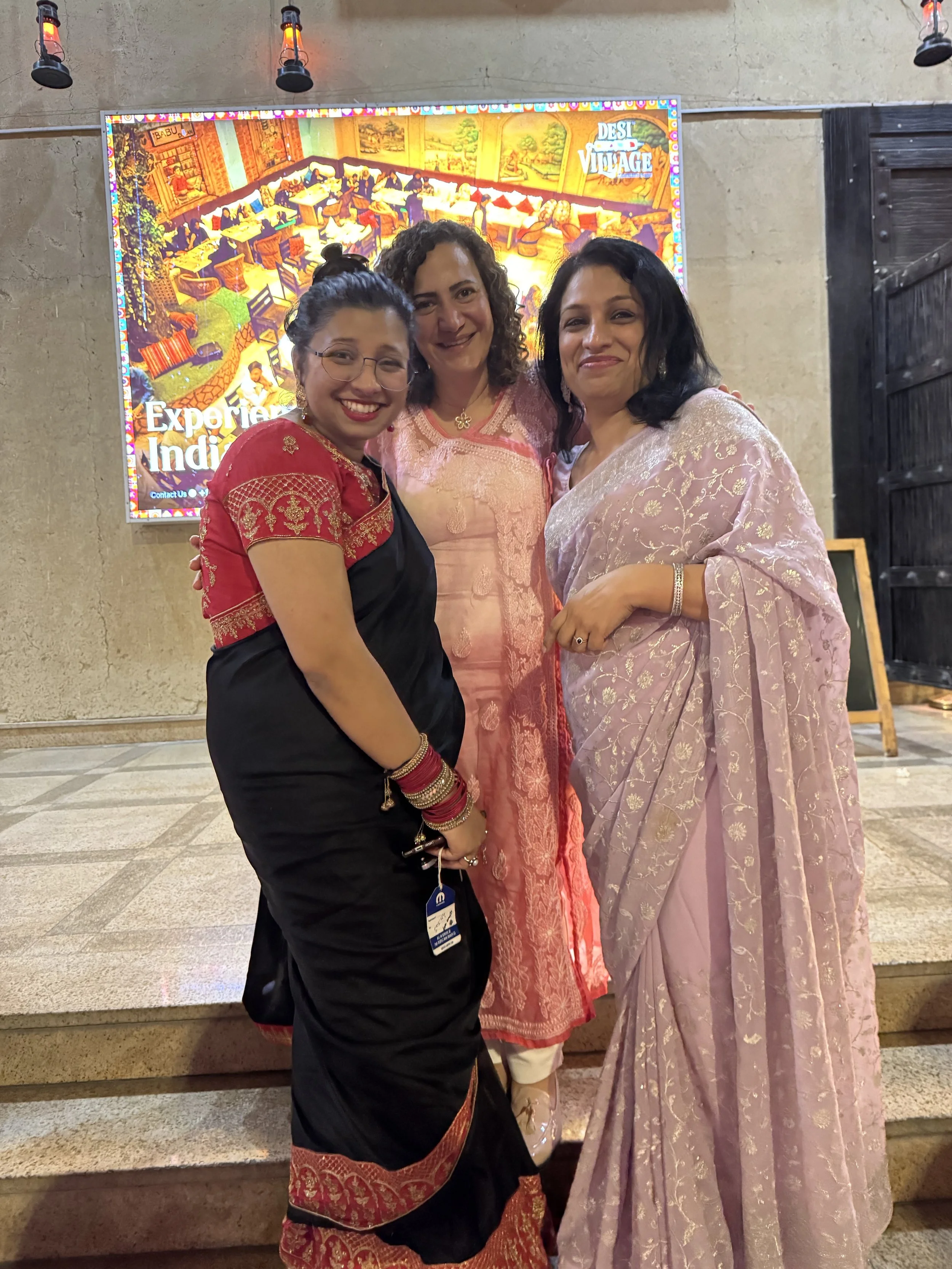 Three women wearing traditional Indian sarees and jewelry, smiling and standing together indoors in front of a colorful image of an Indian village scene on a screen.