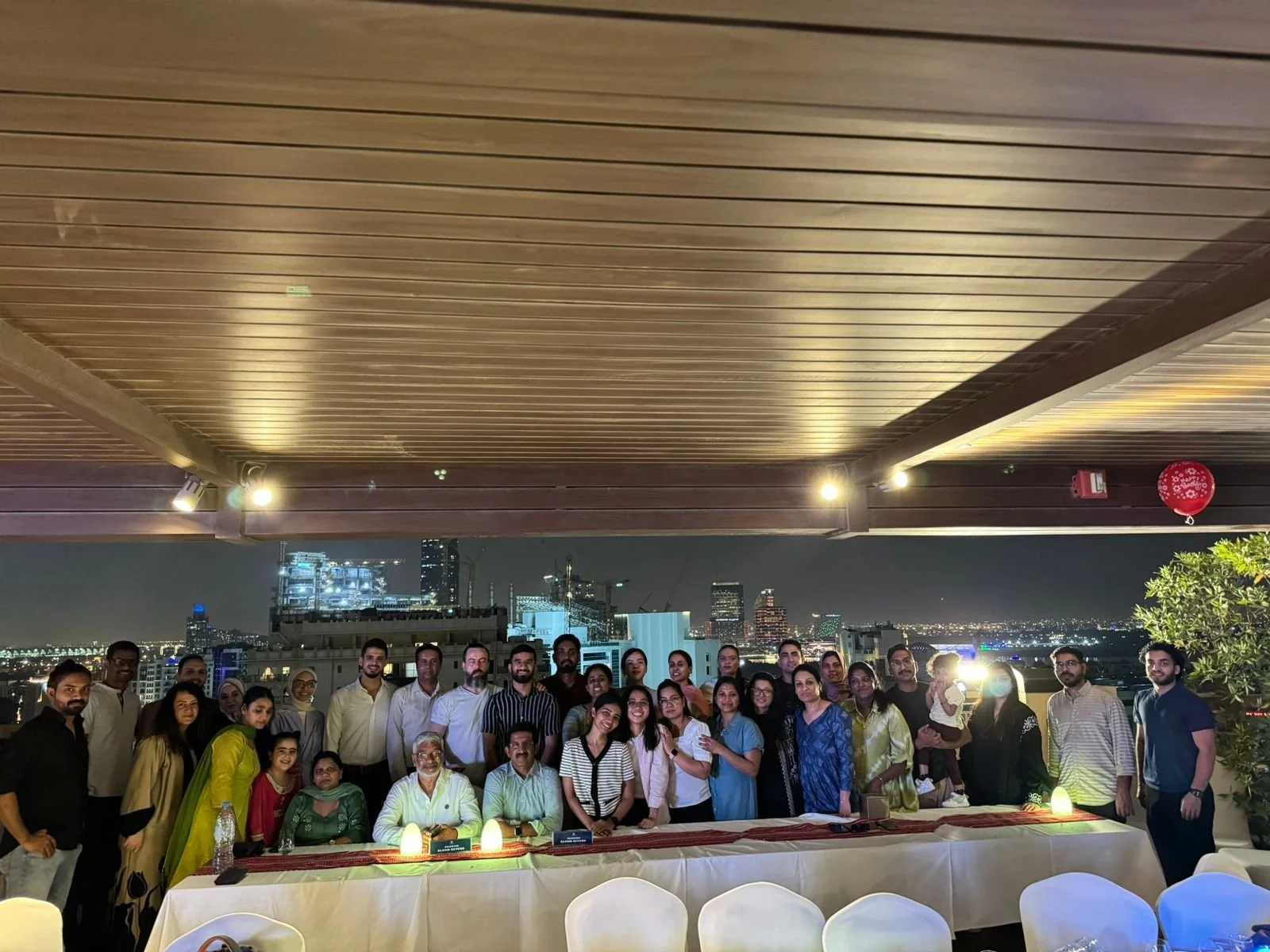 A large group of people gathered on a rooftop at night, with a city skyline in the background, celebrating together at a table with dinner.