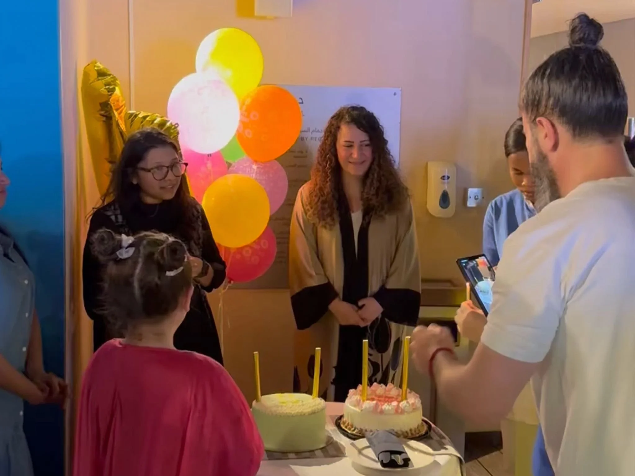 Group of children and adults celebrating a birthday indoors with balloons and cakes.