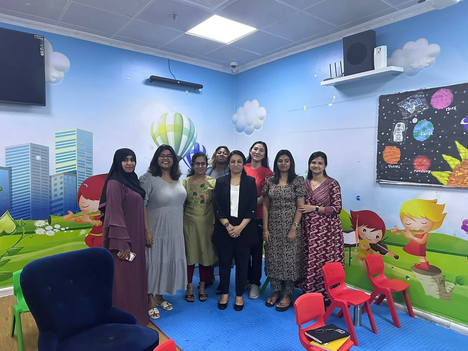 Group of nine women standing in a colorful children’s classroom with painted walls of buildings, clouds, and cartoon children playing instruments.
