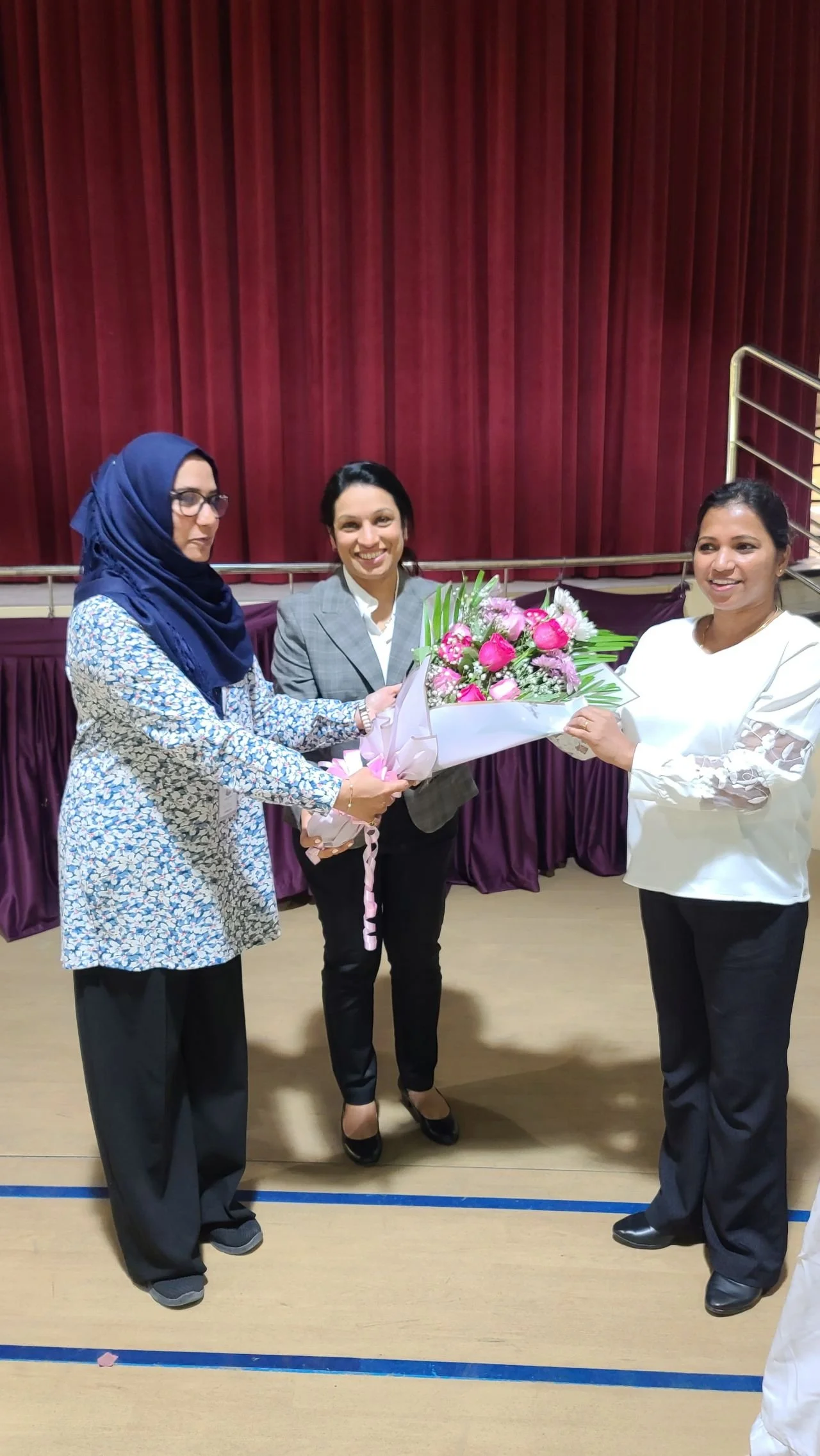 Three women are standing together on a stage, with a red curtain in the background. The woman on the right is handing a bouquet of pink and white flowers to the woman on the left. The woman in the middle is smiling and holding the flowers.