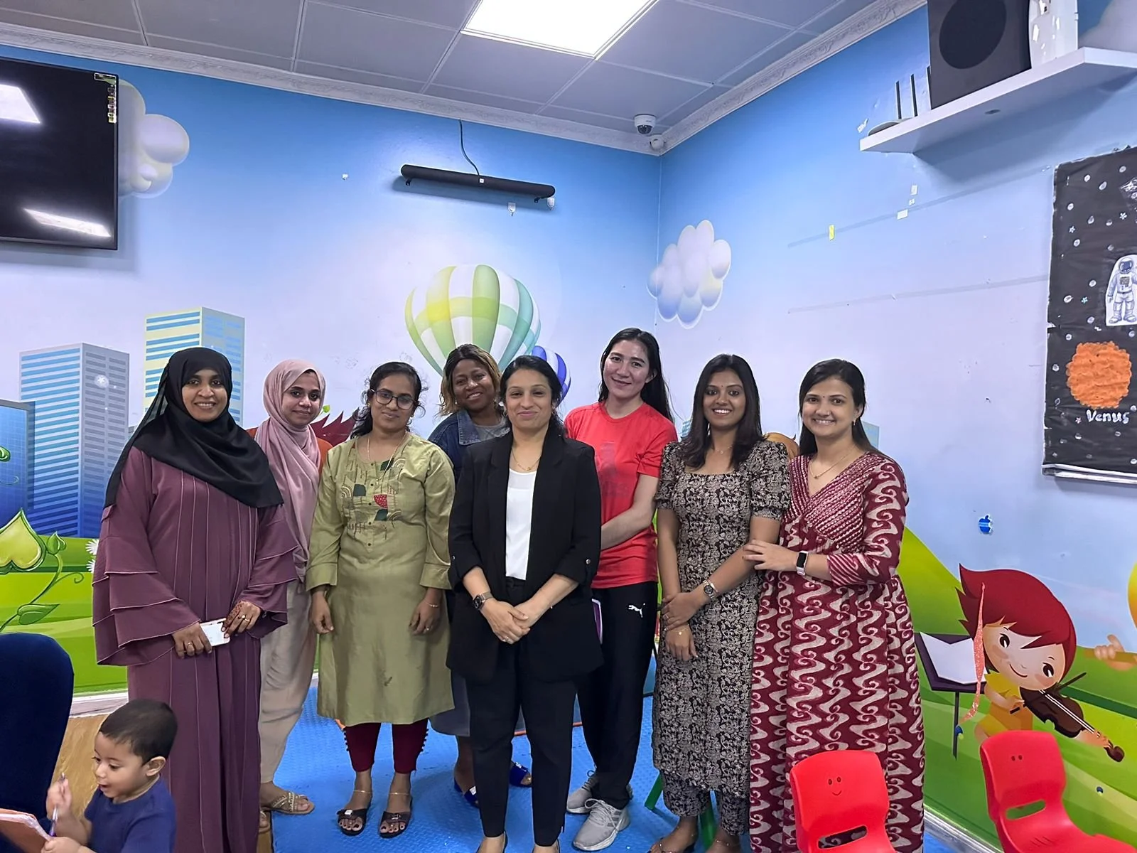 Group of eight diverse women standing together indoors in front of a colorful, cartoon-themed wall, smiling at the camera. One child is in the foreground.