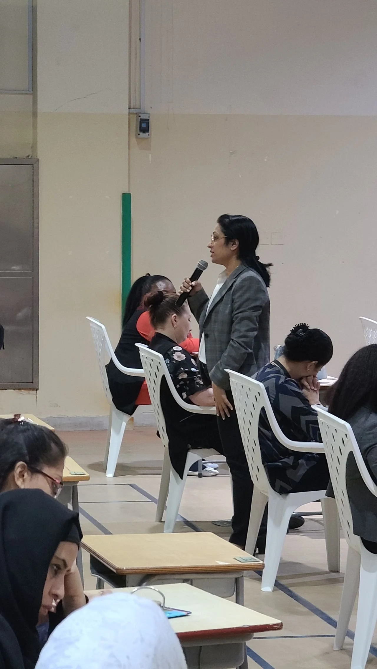 A woman in a gray blazer speaking into a microphone while standing amongst seated women during a meeting or conference.