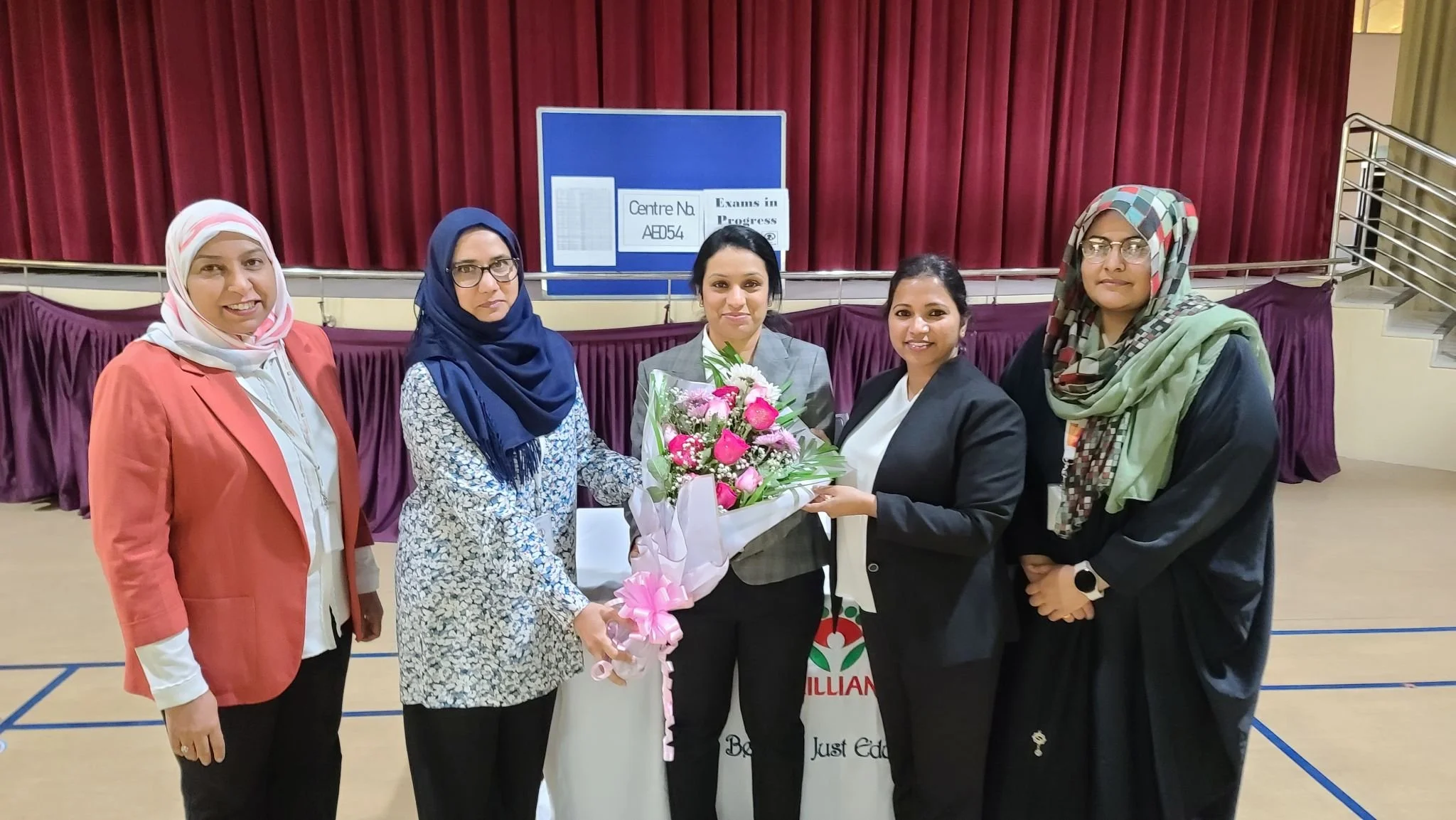 Five women standing together in a gymnasium, holding a bouquet of pink and white flowers, with a red curtain and a blue sign in the background.