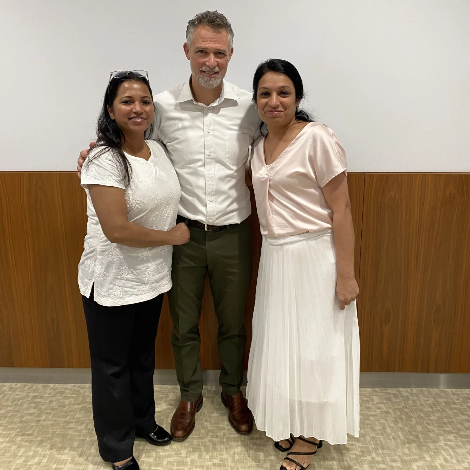 Three people standing together indoors near a wooden wall, smiling at the camera. The person on the left is a woman with long black hair wearing glasses on her head, a white short-sleeved blouse, black pants, and black shoes. The person in the middle is a tall man with gray hair and beard, wearing a white collared shirt, olive green pants, and brown shoes. The person on the right is a woman with long dark hair, wearing a light pink satin top and a long white pleated skirt with black strappy sandals.