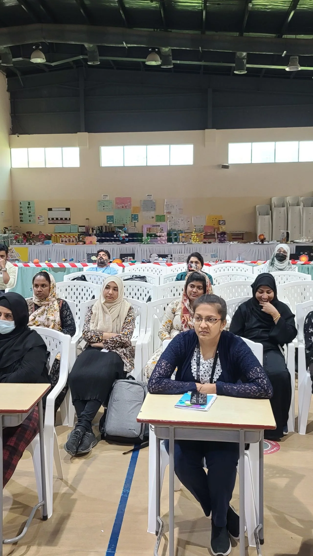 People sitting in chairs inside a large indoor space, possibly a gymnasium or auditorium, with some wearing masks, and a table with various items and displays in the background.