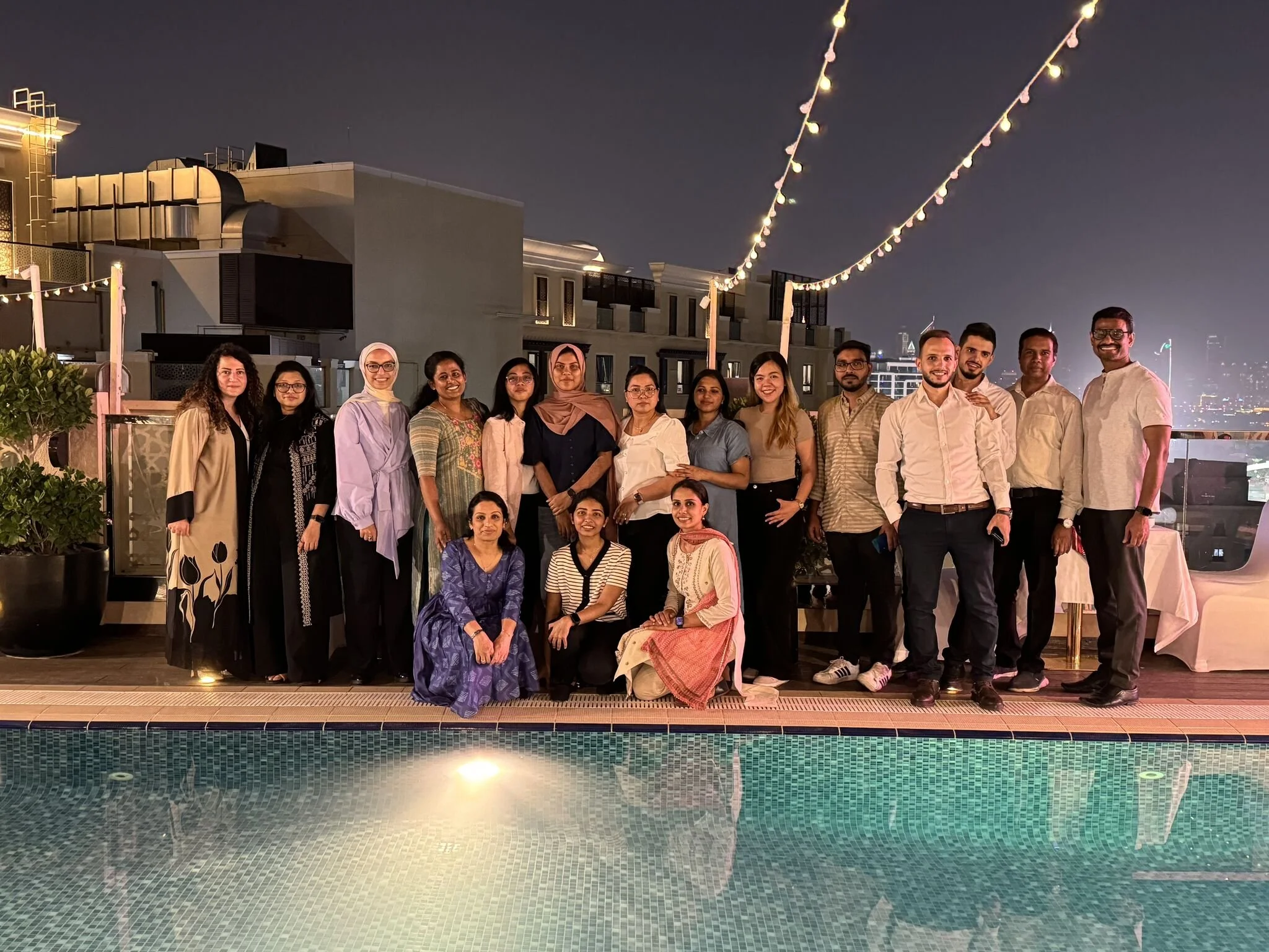 A group of 16 people posing by a pool at night on a rooftop, with city lights in the background and string lights overhead.