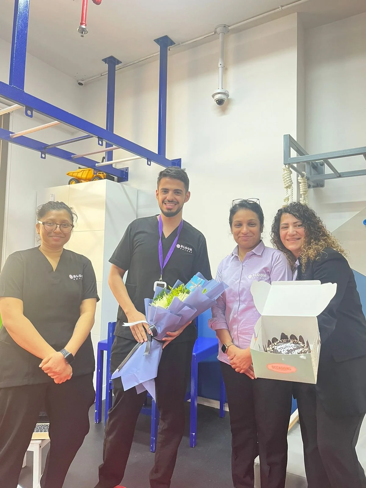 Four people standing together, celebrating with flowers and a cake, in an industrial or restaurant kitchen setting.
