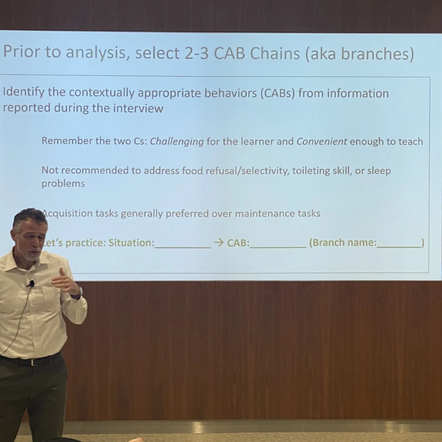 A man giving a presentation in front of a projected slide titled 'Prior to analysis, select 2–3 CAB Chains (aka branches)'. The slide contains instructions and guidelines for selecting behaviors for interview analysis, with a section for practice situation, CAB, and branch name.