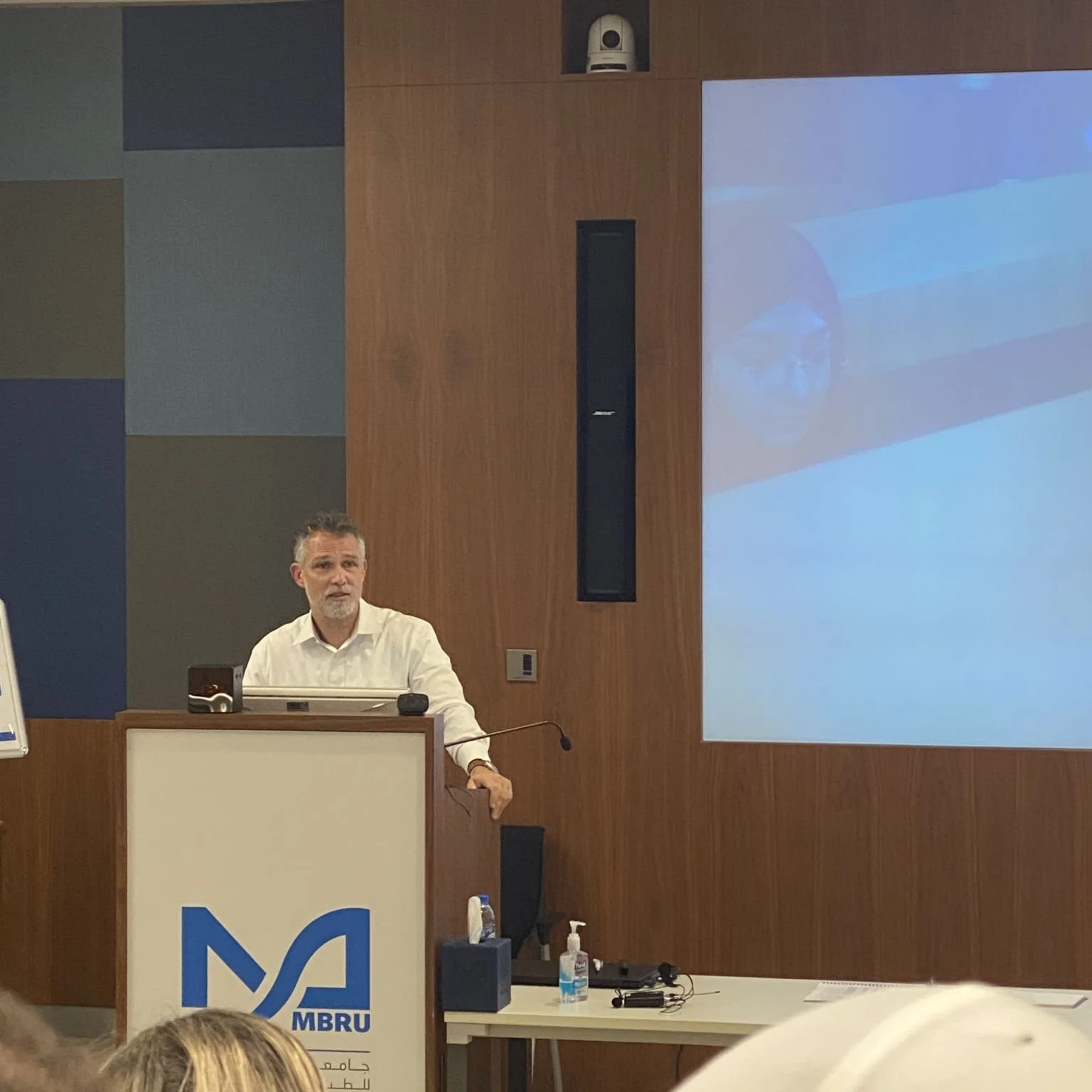 A man presenting at a conference or lecture, standing behind a podium with a logo reading 'MBRU' and a large screen displaying a projected image.