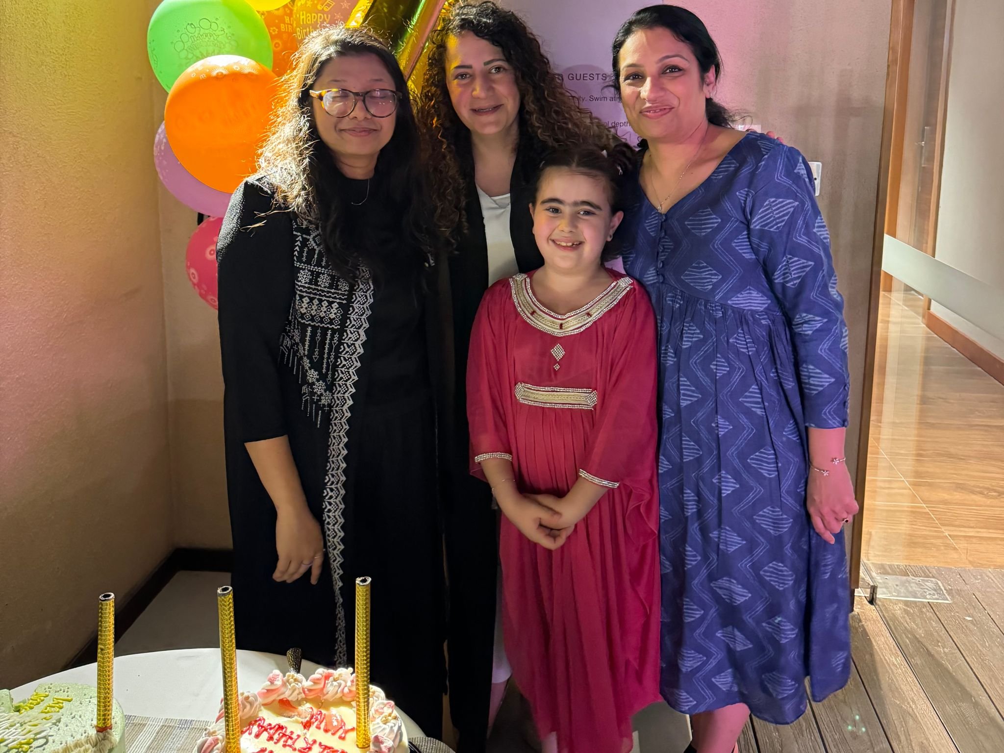 Four women and a girl smiling at a birthday party, with balloons and birthday cake on the table in front of them.