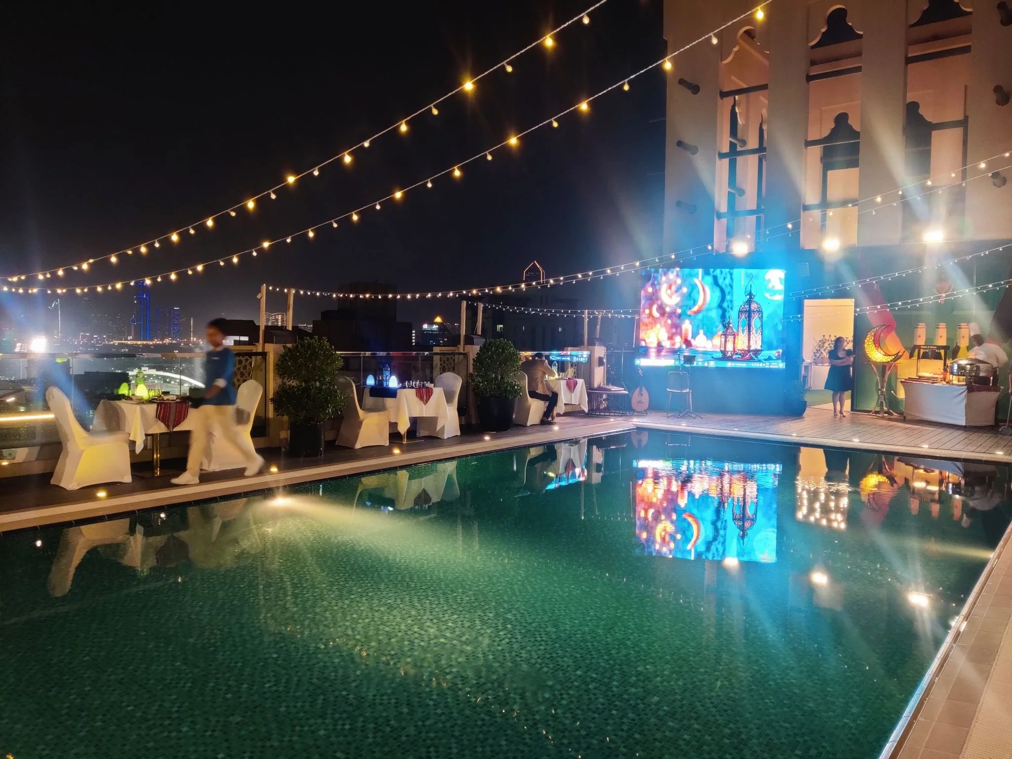 Outdoor rooftop party with string lights, dining tables, a swimming pool, and city skyline at night, featuring a large digital screen and decorations.