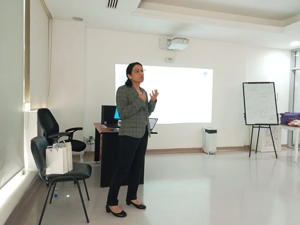 A woman with glasses presenting in a bright, modern conference room with a whiteboard, projector, and chairs.