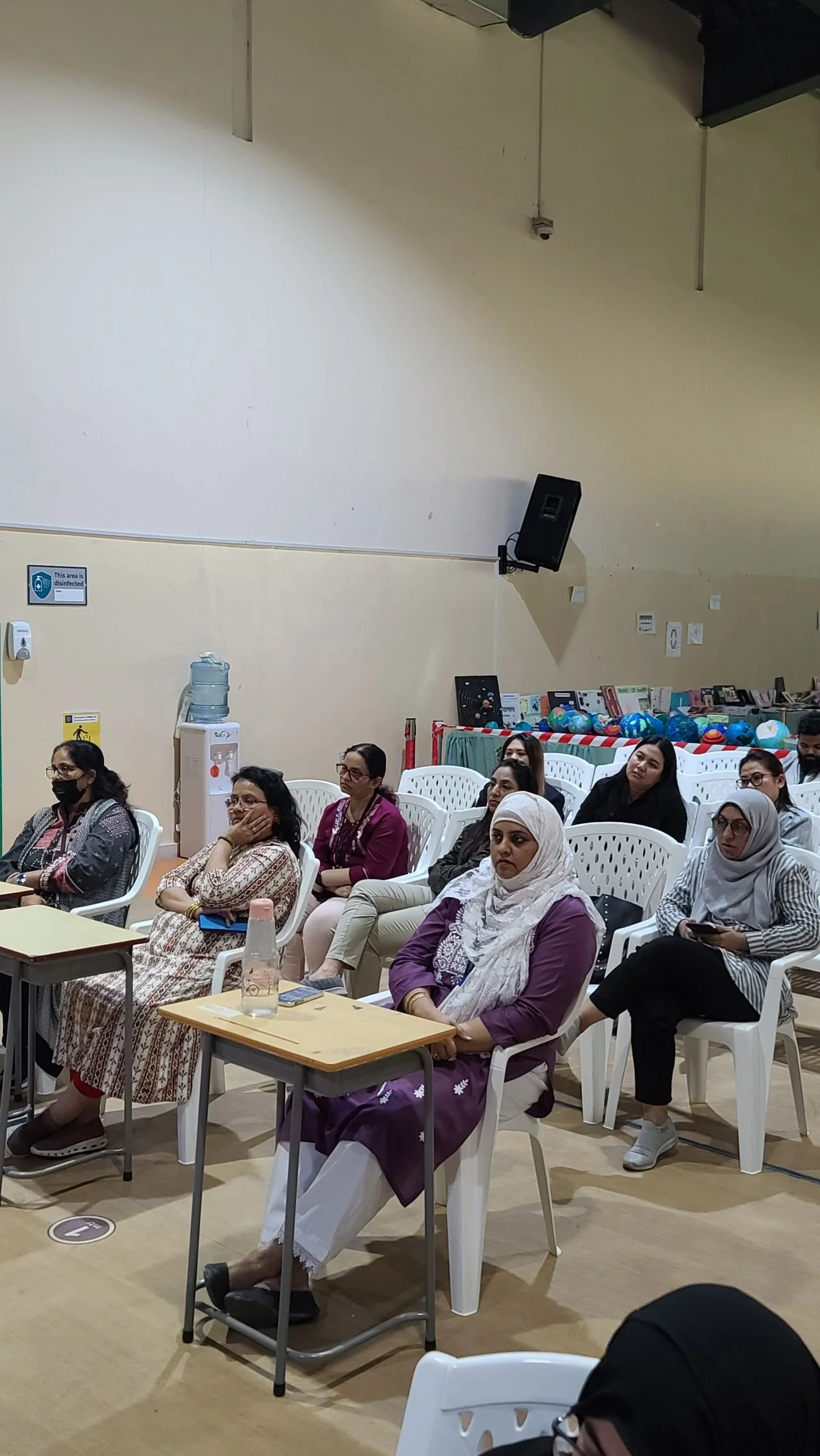 A group of women sitting in white plastic chairs in a room, some listening attentively and some using mobile devices, with a wall, water dispenser, and a display of colorful balls in the background.