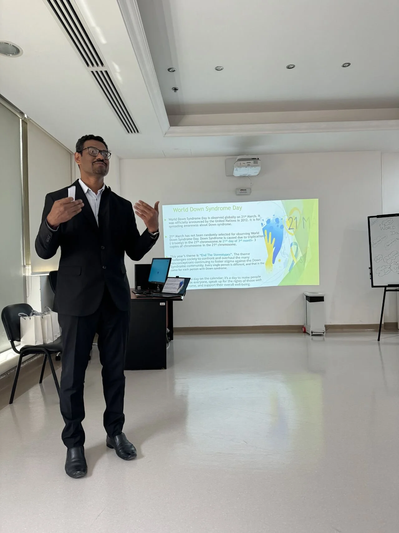 A man in a black suit, white shirt, and glasses standing in front of a presentation slide about World Down Syndrome Day in a conference room. The slide discusses the significance of March 21st and Down syndrome awareness.