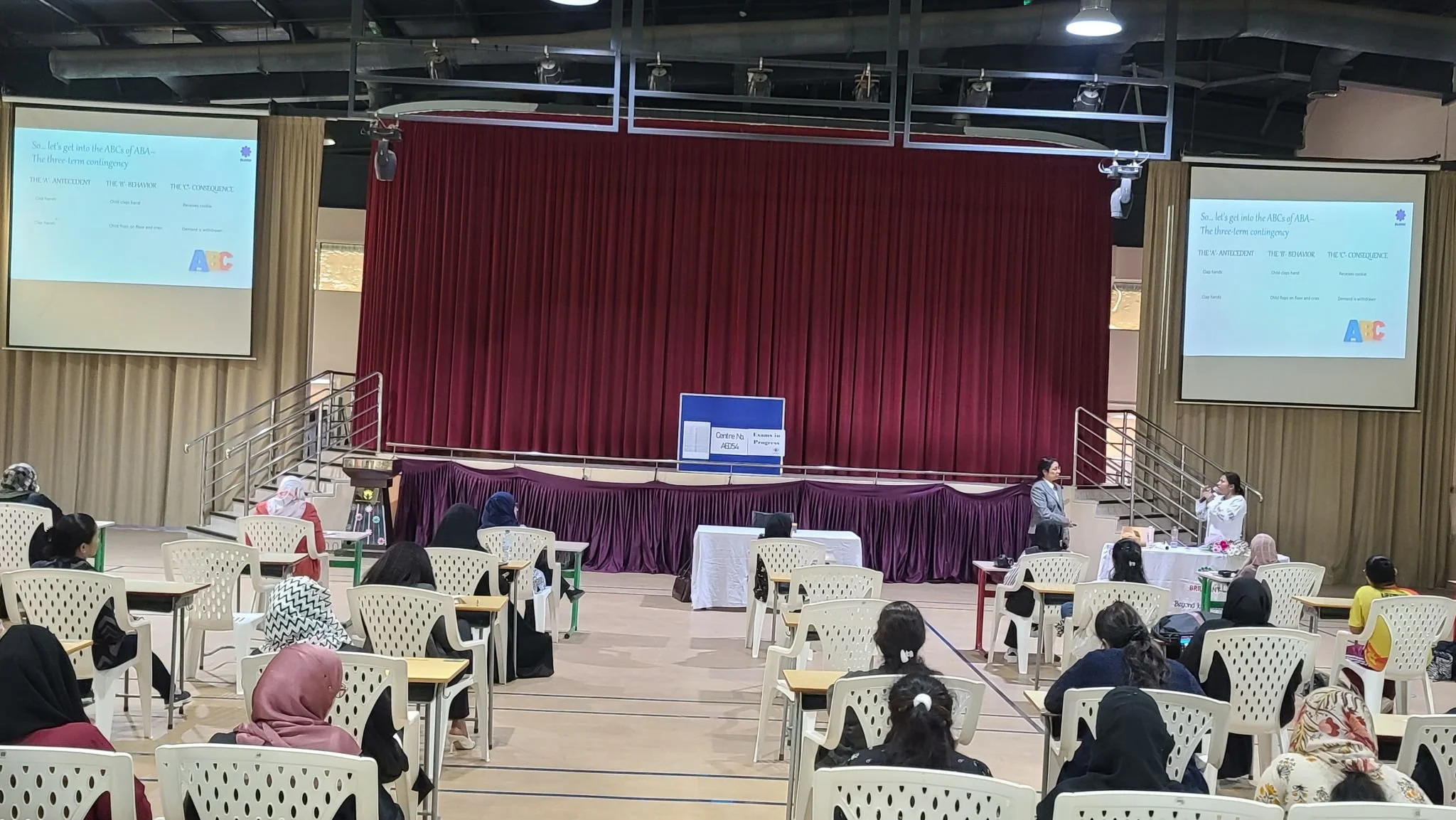 A classroom or seminar room with an audience seated at desks, facing a stage with red curtains. Two presenters, a man and a woman, are standing on the stage in front of the curtains, speaking into microphones. Two large screens display a presentation about ABCs of ABA.
