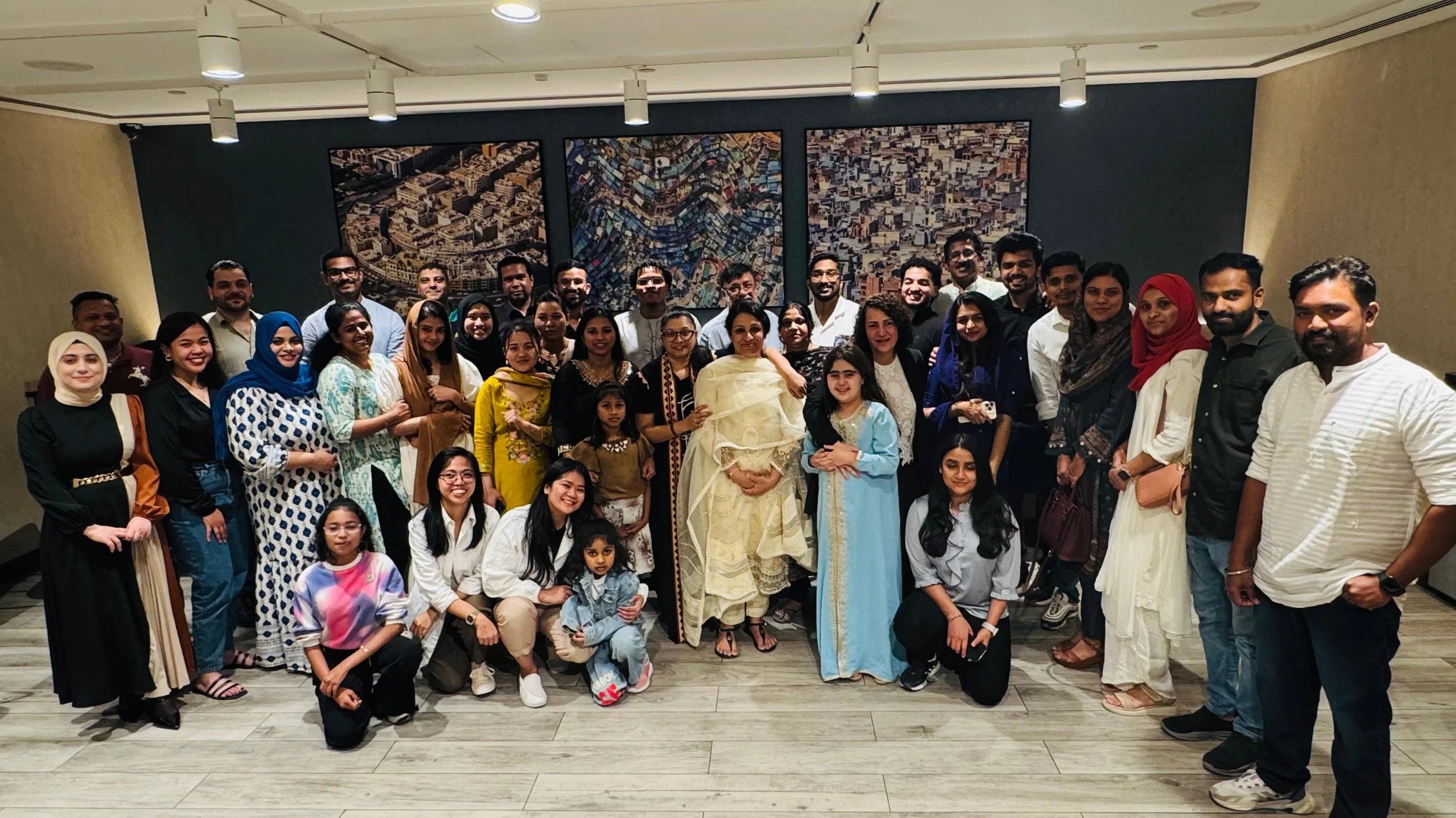 Group of people standing together for a photo in an indoor setting. The group includes men, women, and children of diverse backgrounds, with some wearing traditional South Asian attire and others in casual clothing. The background features three large aerial cityscape paintings or photographs on the wall.