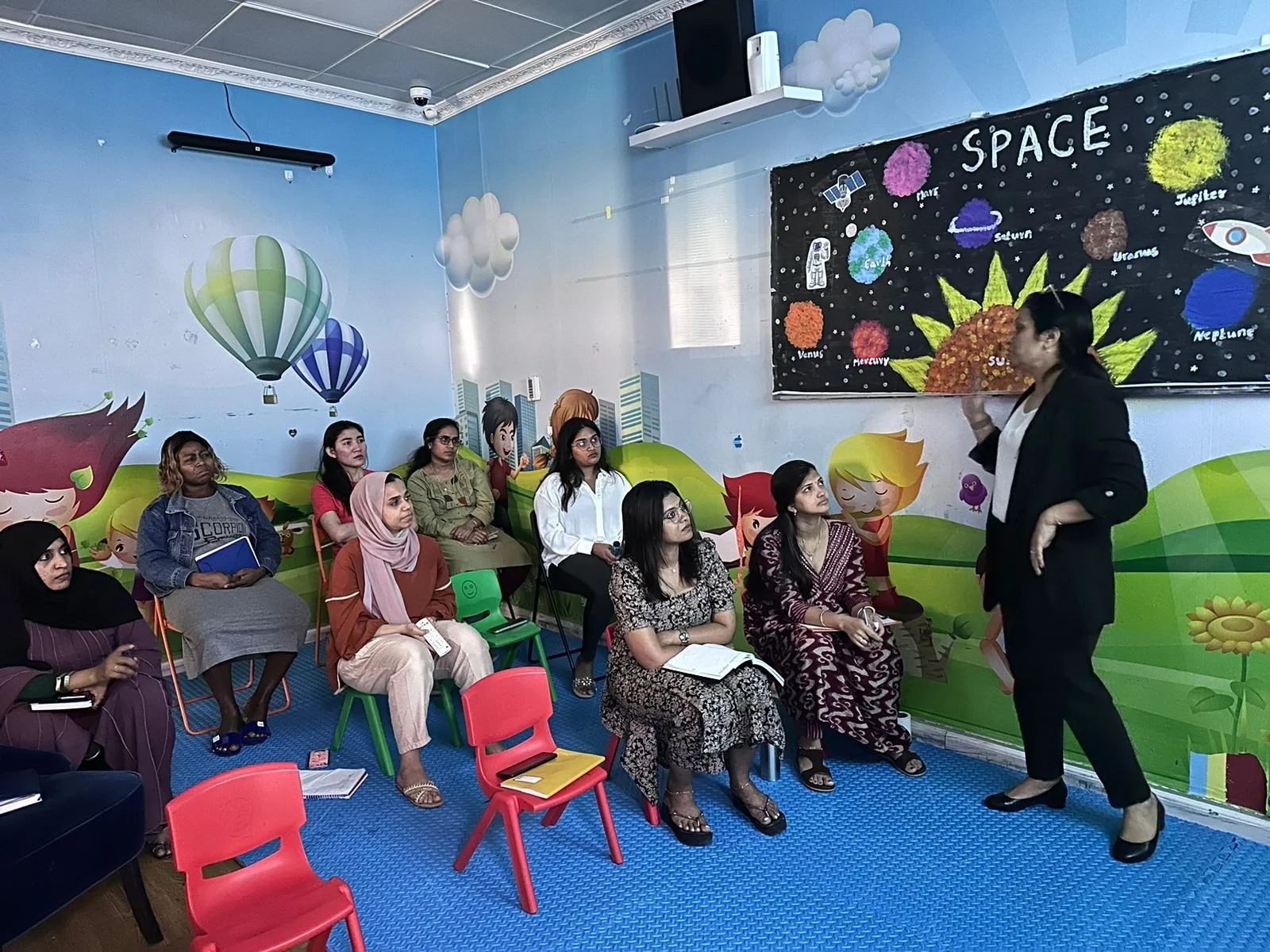 A woman in black talking to a group of women seated in colorful chairs in a classroom decorated with space-themed mural and wall celebrating space planets