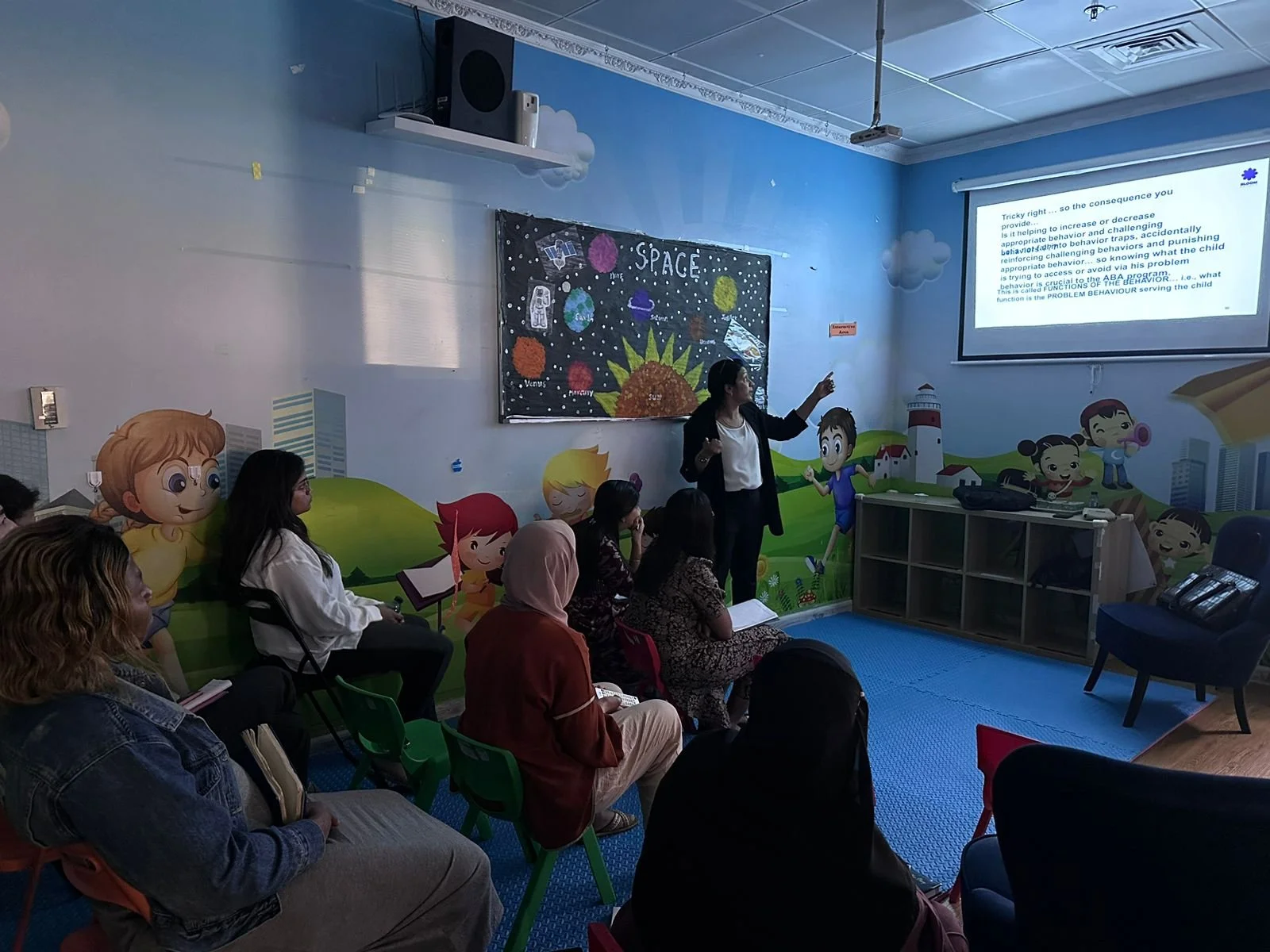 A classroom with a teacher presenting a slide to a group of adults, with colorful space-themed murals and cartoon characters on the walls.