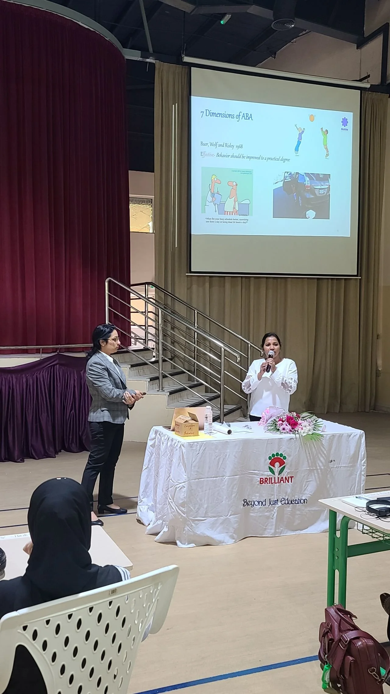 A presentation in a large room with a speaker and a woman standing near a table. The table has a floral arrangement and a banner with the words 'BRILLIANT Beyond Just Education'. A projector screen displays a slide titled '7 Dimensions of ABA' with images and text.