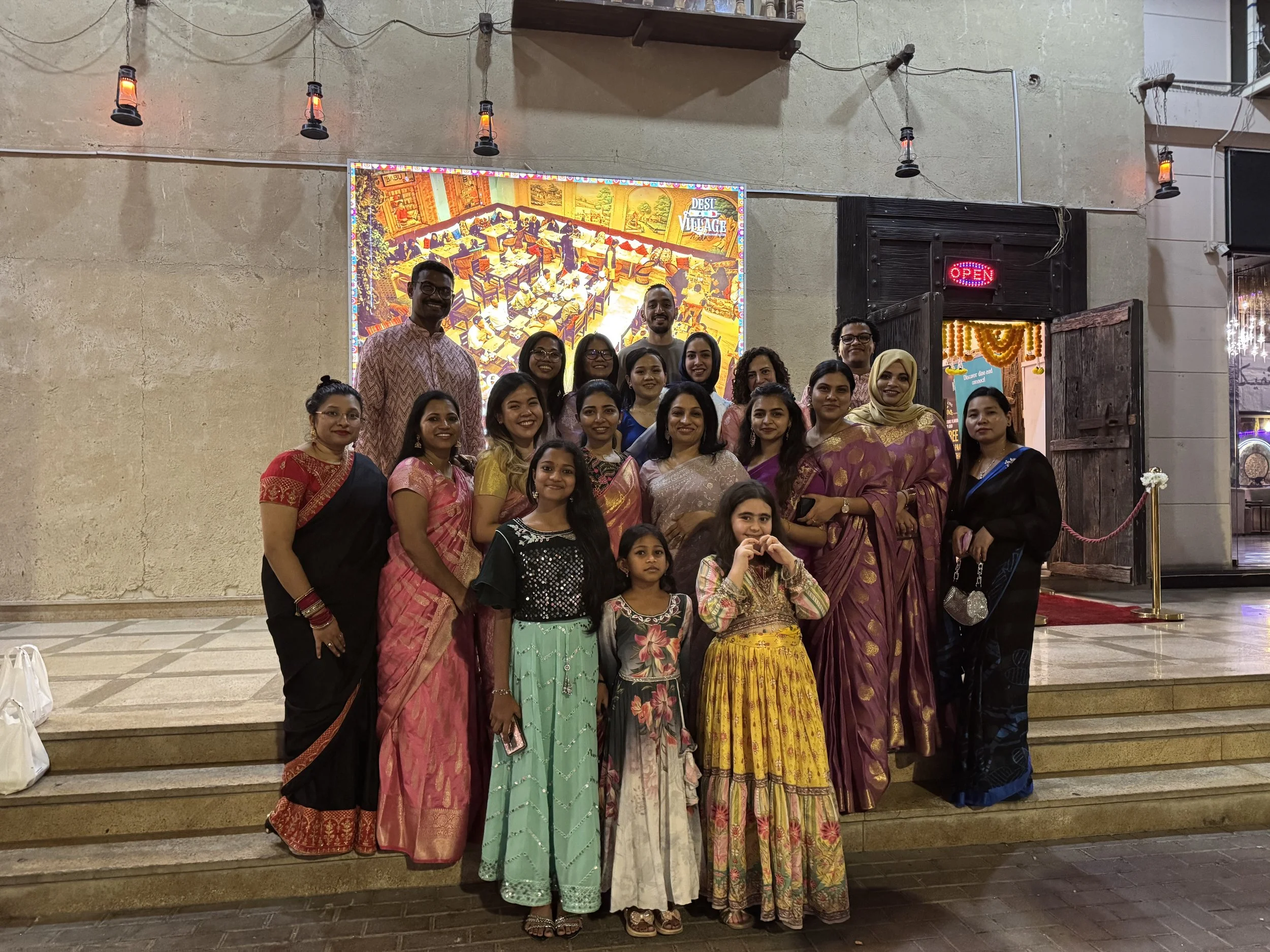 Group of people dressed in colorful traditional Indian attire, standing on steps outside of a restaurant with a neon 'Open' sign, a large digital screen displaying an indoor scene in the background, and a doorway decorated with flowers.