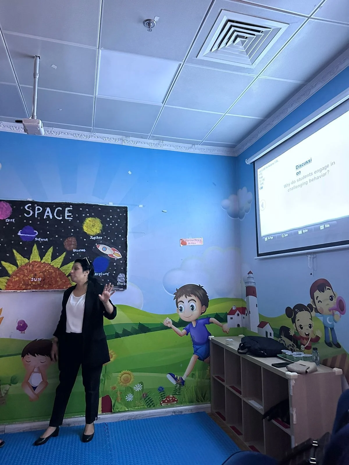 A woman giving a presentation in a classroom with a colorful space-themed mural and a projection screen displaying a question about challenging behavior.
