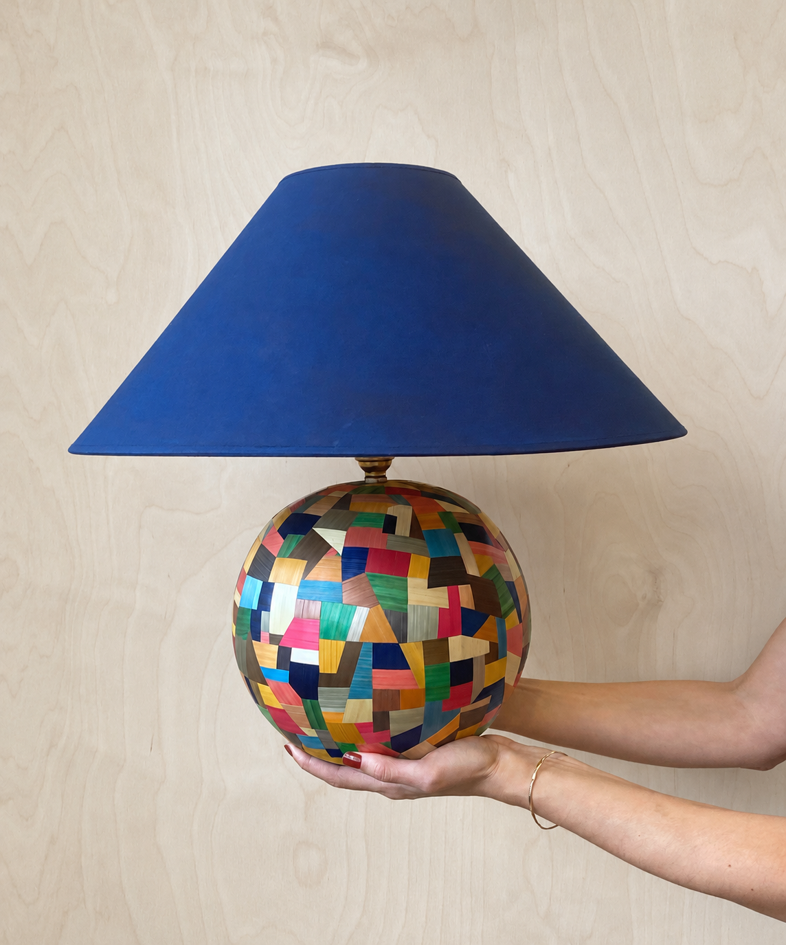 A colourful spherical straw marquetry table lamp by Eleanor Nadimi, with a patchwork of geometric shapes and a deep blue conical lampshade.