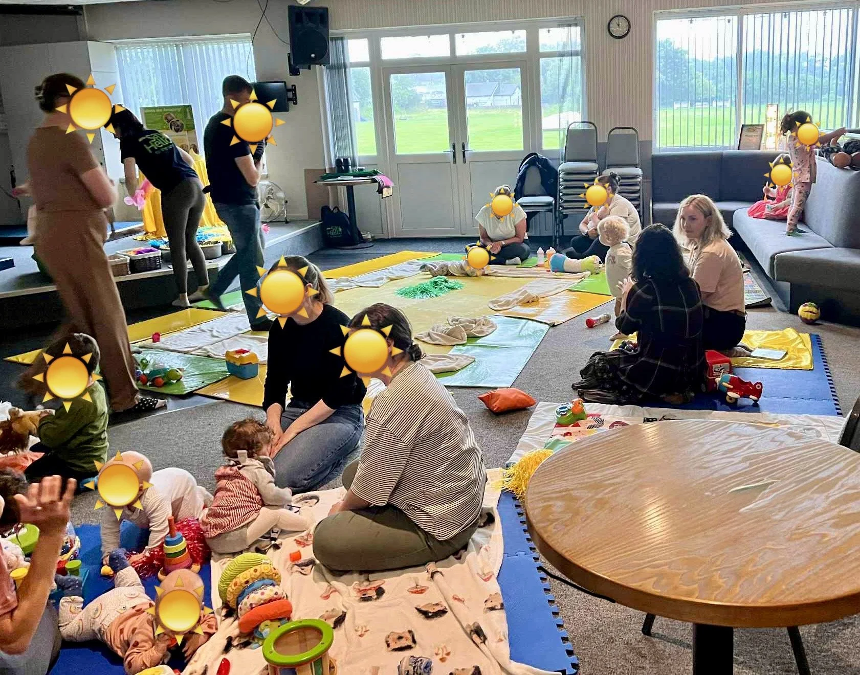 Children and adults participating in a music or play session in a bright room with large windows, mats on the floor, toys, and some chairs and playing areas.
