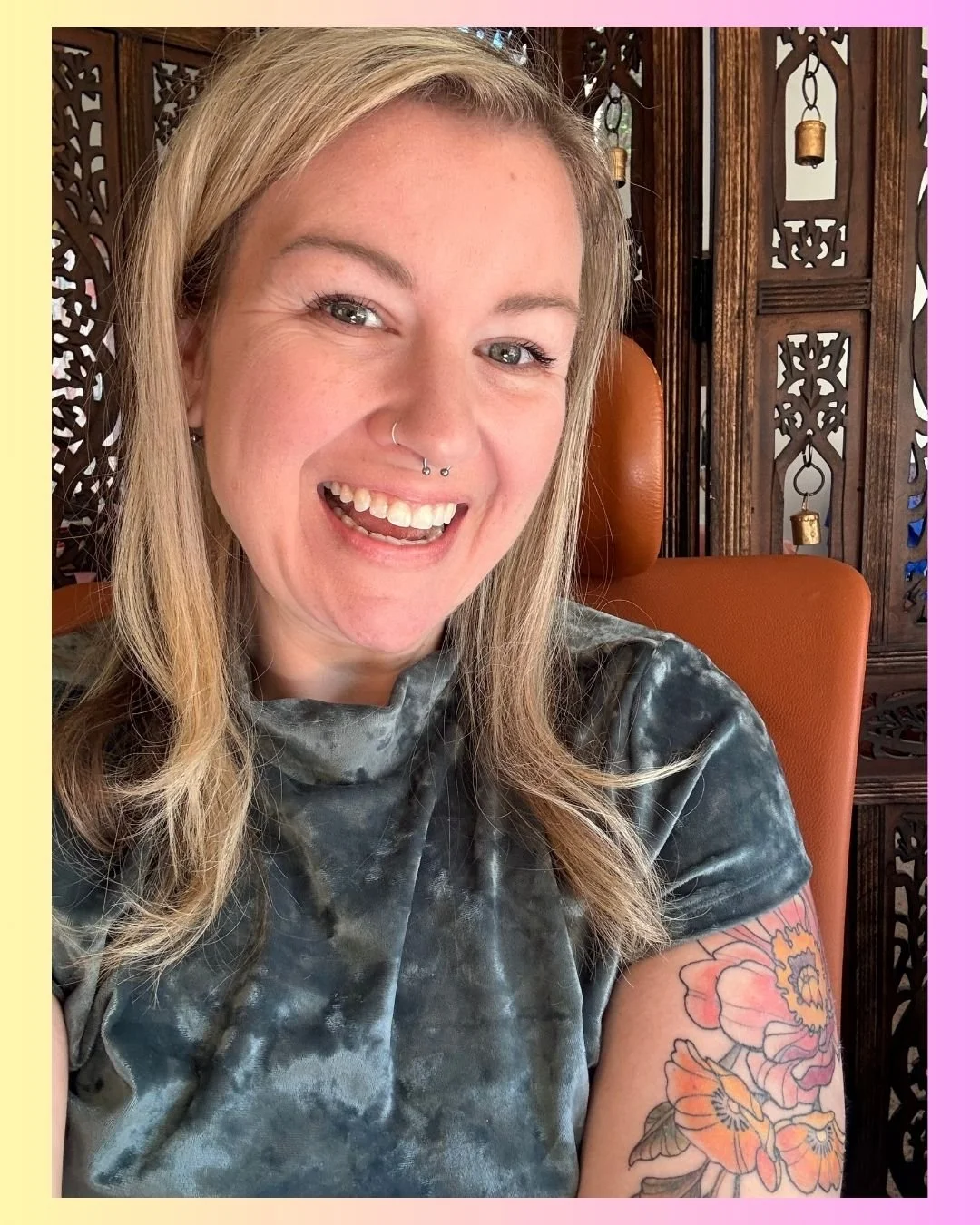 A woman with blonde hair smiling, showing her teeth, and wearing a tie-dye shirt. She has a nose piercing and an arm tattoo of colorful flowers. She is sitting on a brown leather chair in front of a wooden decorative screen with hanging bells.